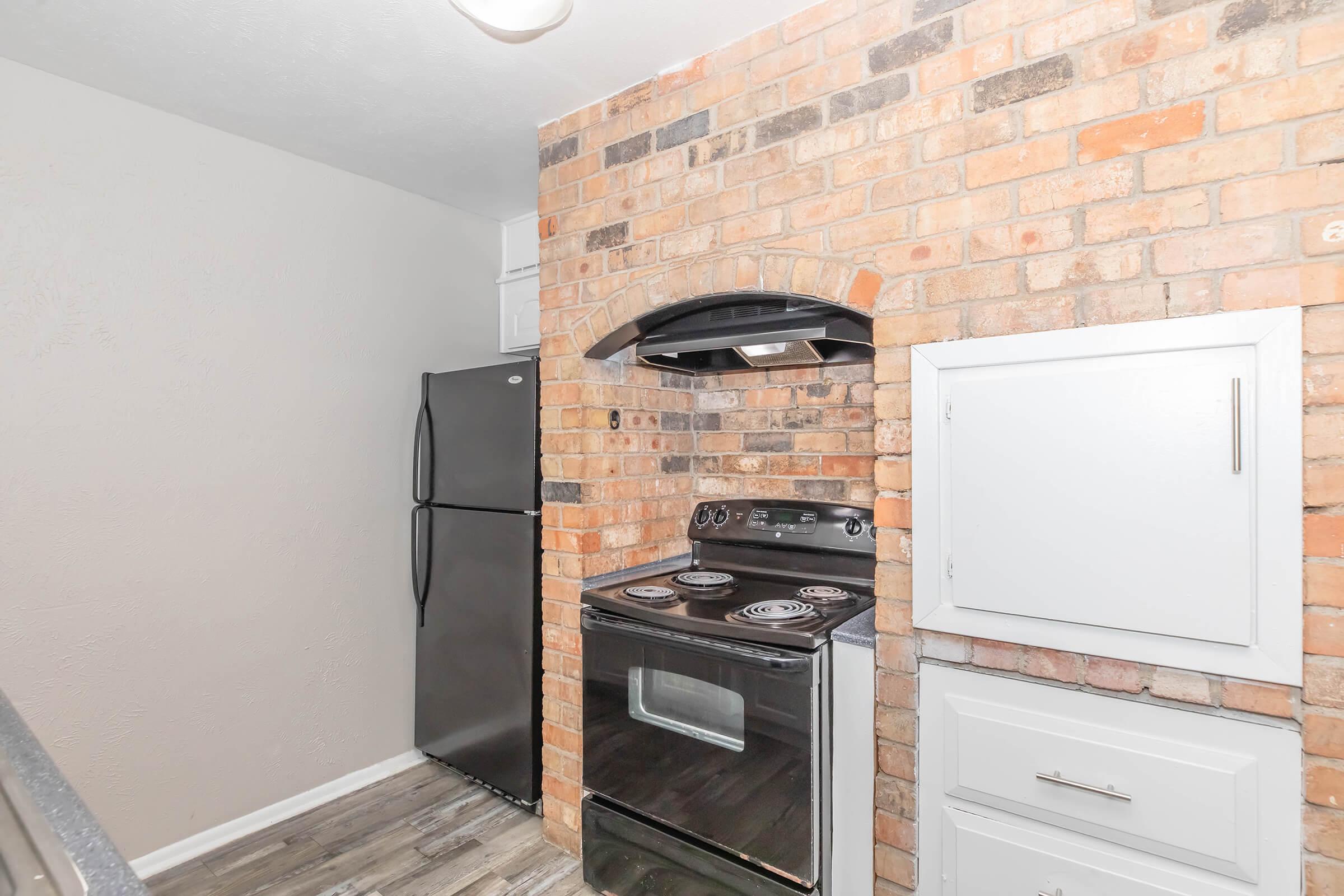 A small kitchen featuring a black refrigerator, an oven with a ceramic cooktop, a brick accent wall, and white cabinetry. The kitchen has an open layout with a focus on the stove and hood. The flooring is a modern design, complementing the rustic elements of the brick wall.