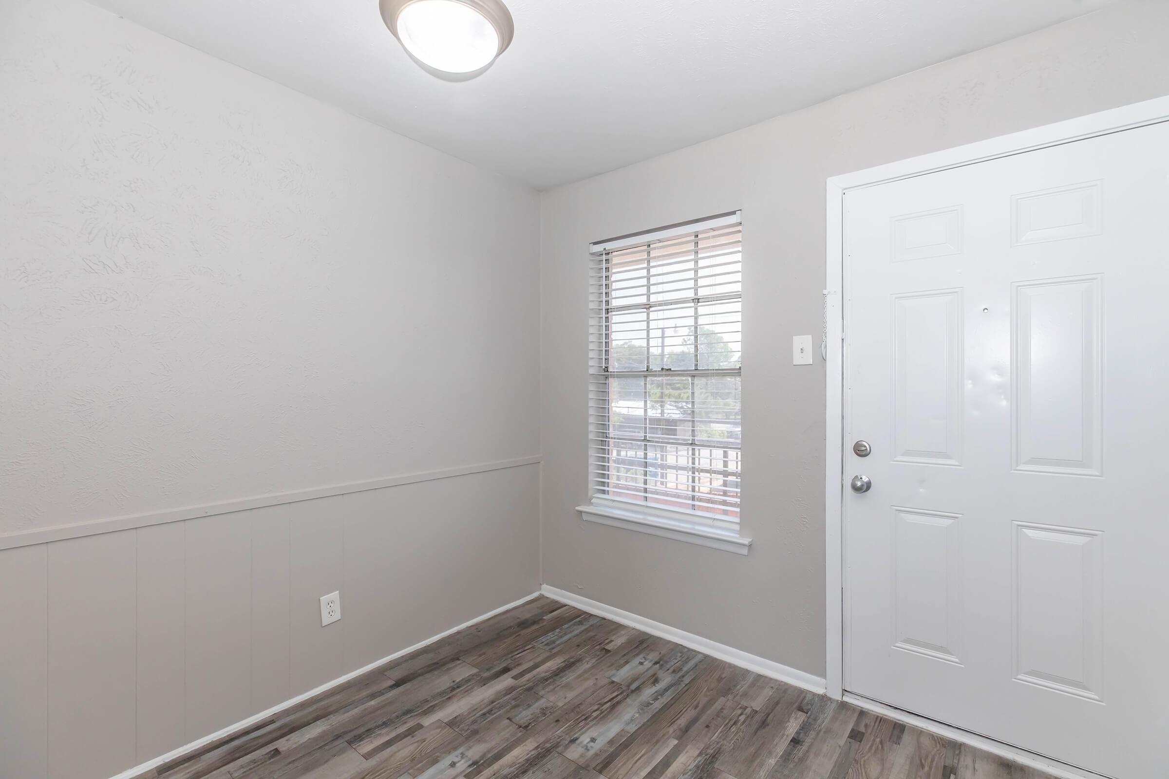A small, well-lit entryway featuring light gray walls, a window with white blinds, and wood-like laminate flooring. A white door with a doorknob is visible on the right, and a ceiling light fixture hangs above. The space is minimalistic and inviting.