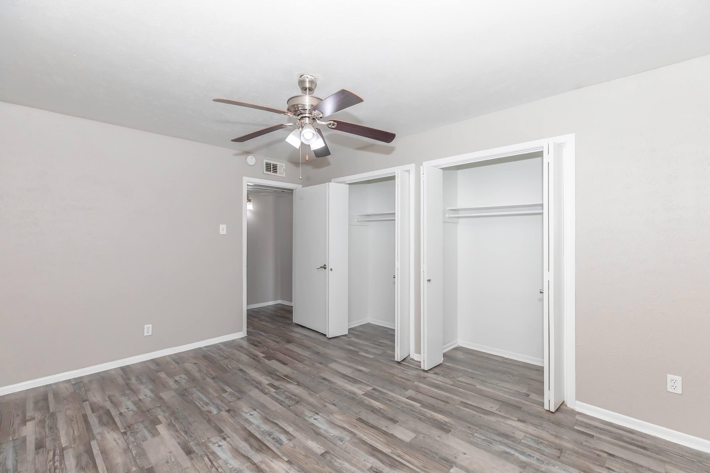 A spacious, empty room with light gray walls and wooden laminate flooring. It features a ceiling fan with five blades, two open white closet doors, and a doorway leading to another room in the background. The overall atmosphere is bright and uncluttered, ideal for decoration or furniture placement.