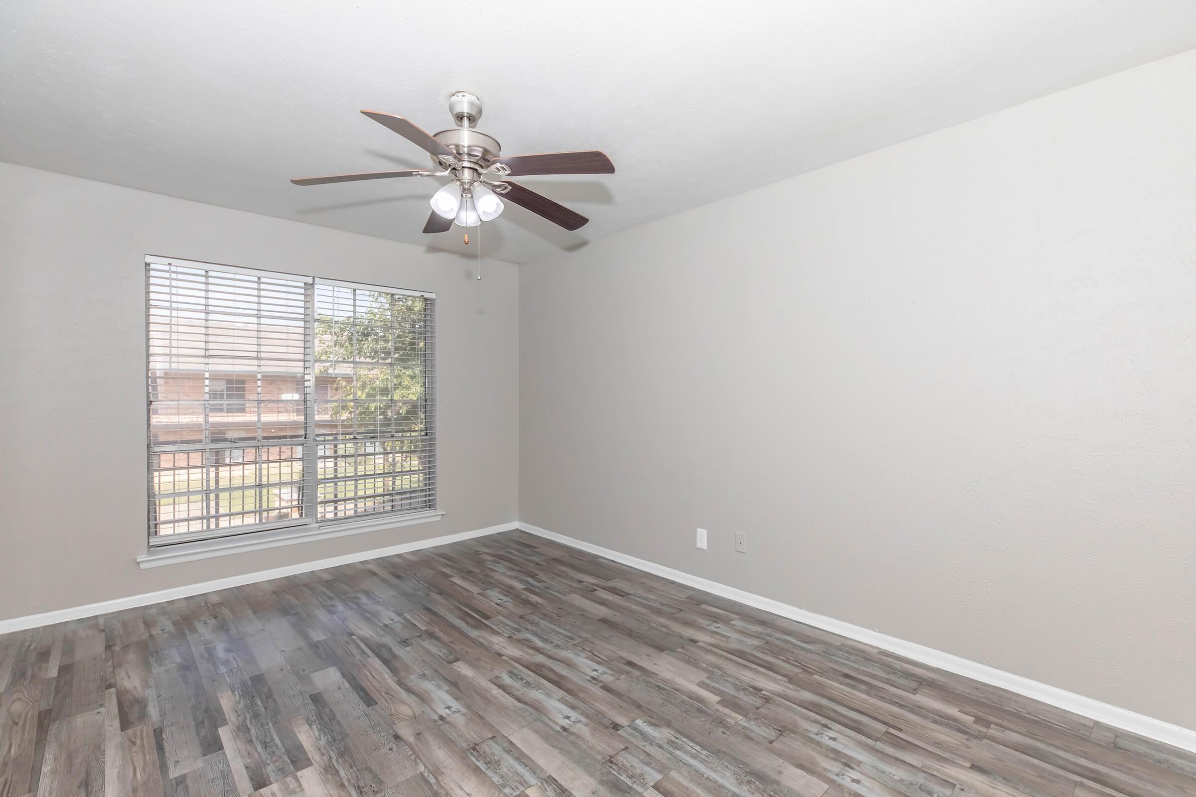 A well-lit, empty room with beige walls and a ceiling fan featuring four blades. The floor is made of light-colored wood planks. A window with white blinds allows natural light to enter, offering a view of an exterior area. The space is clean and ready for furnishing.