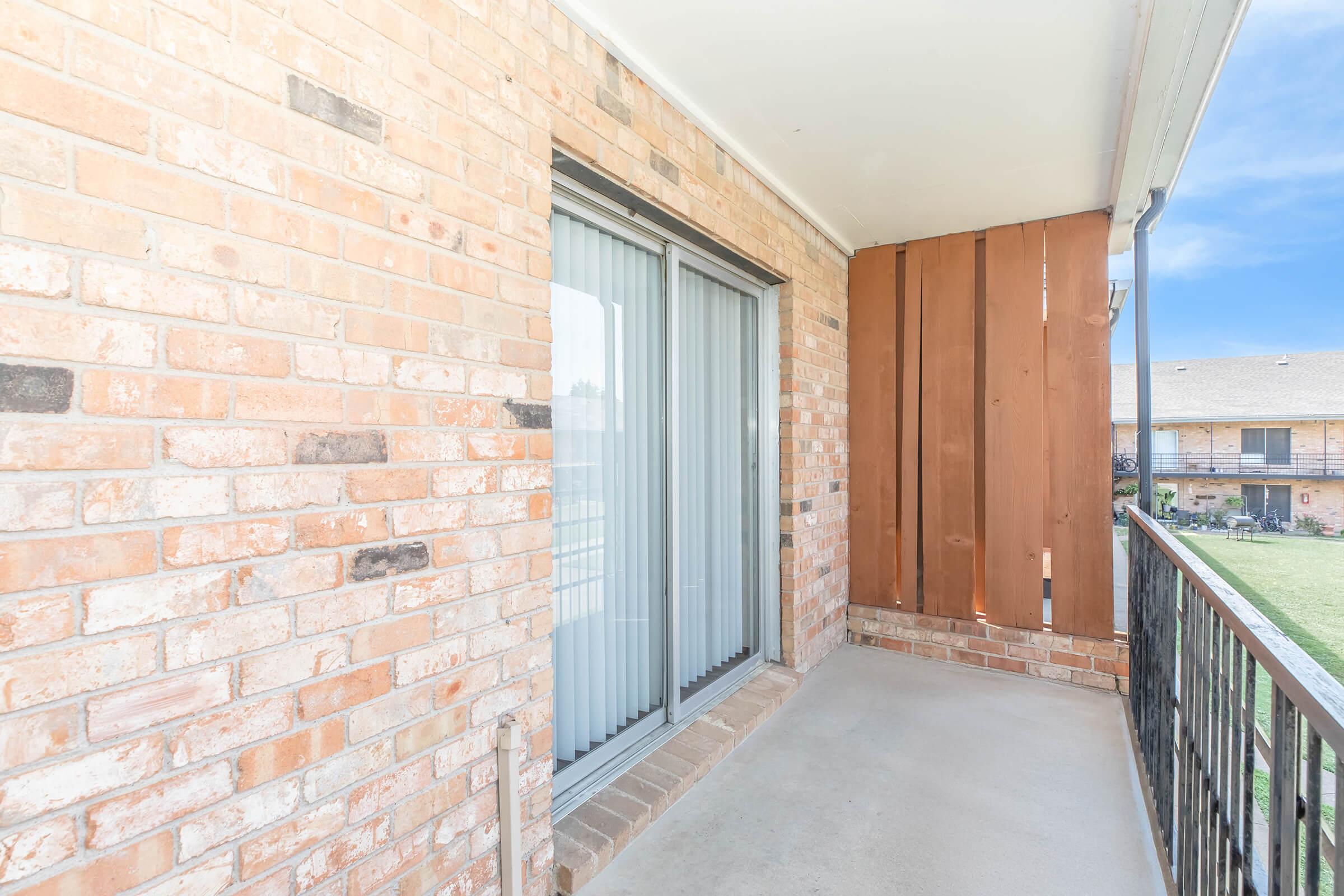 A small balcony with a concrete floor, featuring a sliding glass door and brick walls. One side is adorned with a wooden panel, while the other shows a view of a grassy area outside. The balcony appears clean and well-maintained, with an inviting atmosphere for relaxation.
