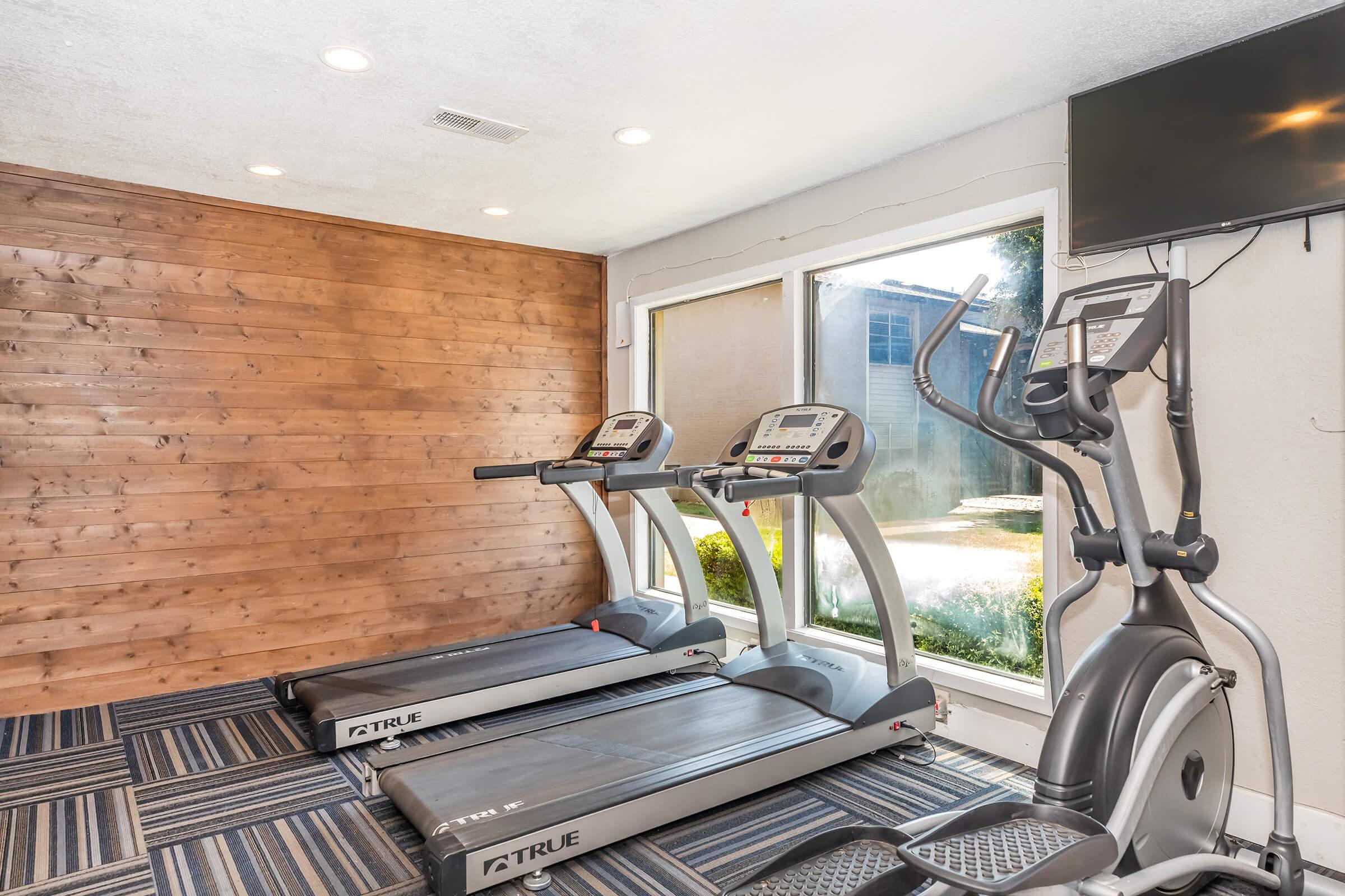 A well-lit gym area featuring two treadmills and an elliptical machine, set against a backdrop of a wooden accent wall and large windows that offer a view of the outside. There is also a mounted television on the wall, and the flooring consists of a patterned carpet.