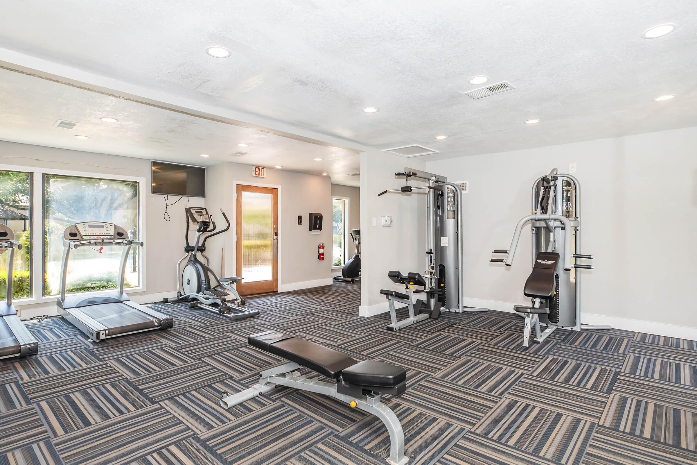 A modern gym interior featuring exercise equipment including treadmills, weight machines, and a workout bench. The space has bright lighting, patterned carpet, and large windows that offer natural light, creating an inviting atmosphere for fitness activities.