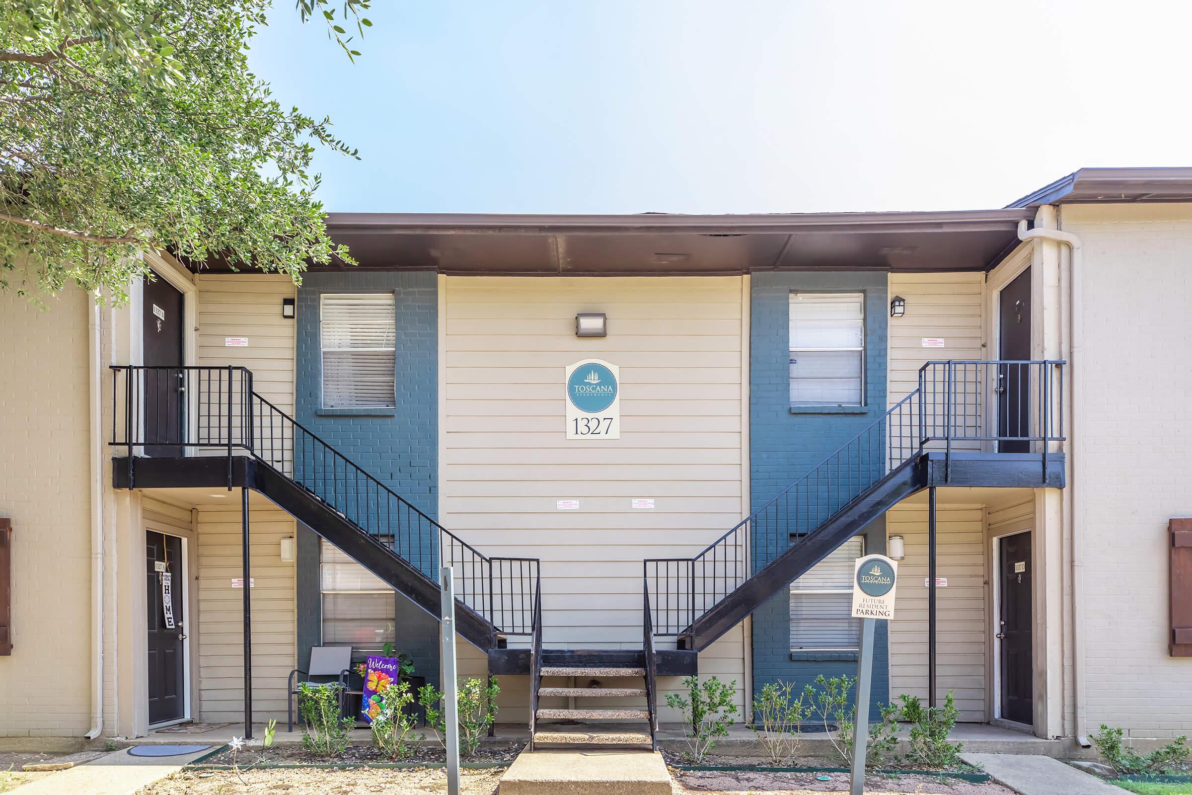 Two-story apartment building with a light-colored exterior and blue accents. The entrance features a set of stairs leading to the second floor. There are two doors on each floor, and a sign displaying the unit number "1327." A small bench is visible in front of the building surrounded by greenery.