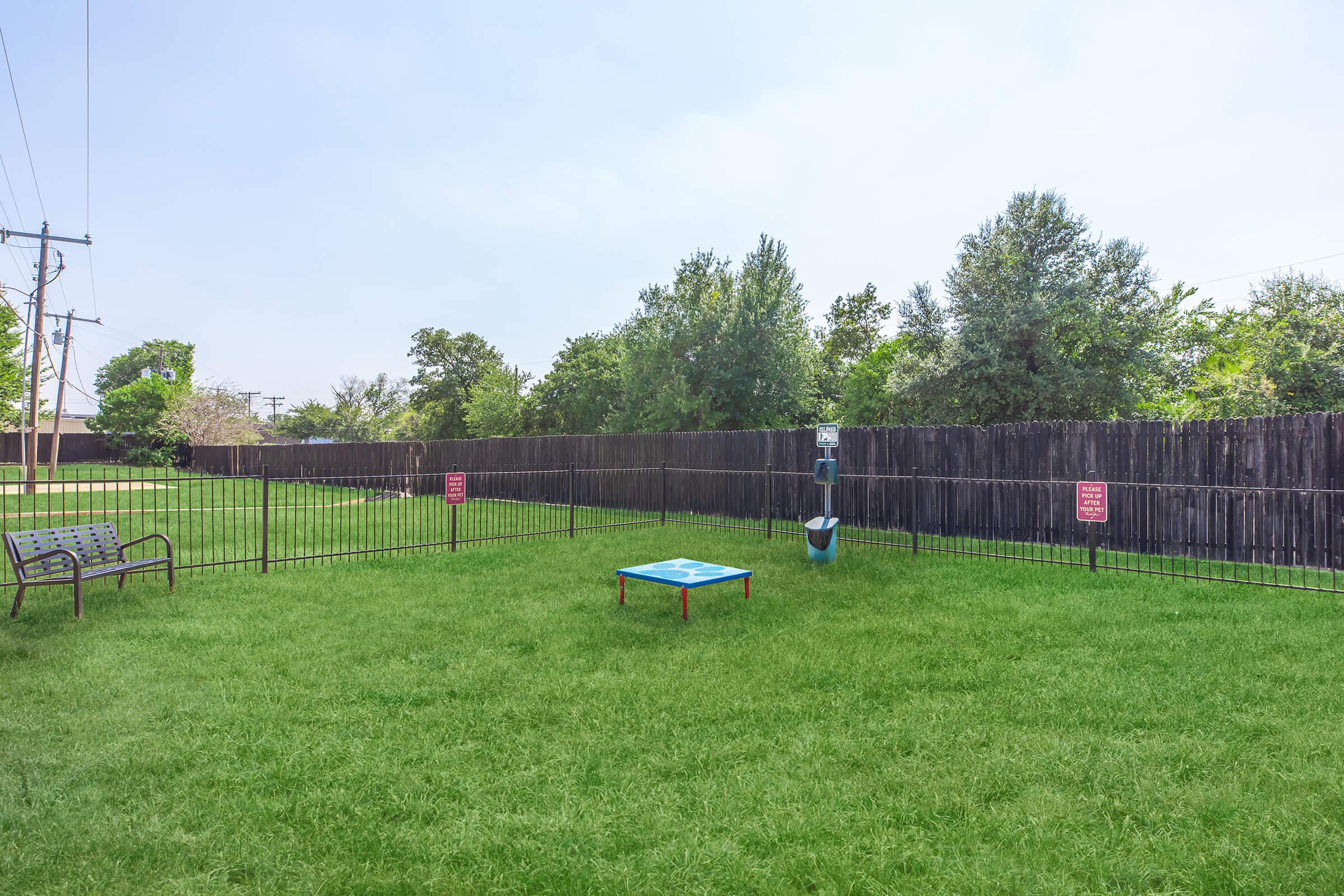 A grassy area enclosed by a black fence, featuring a blue platform in the center and a bench to the side. There are several trees in the background, and signs are visible on the fence. The sky is clear, indicating a bright day.