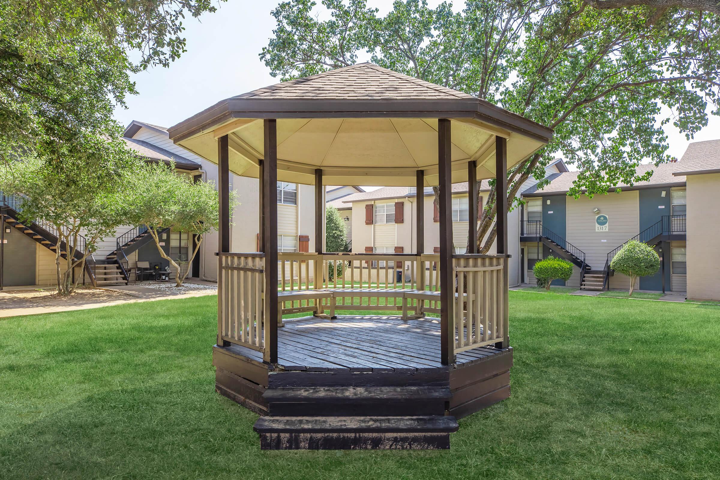 A gazebo with a wooden structure and a pitched roof is situated on a grassy area surrounded by trees. In the background, several apartment buildings are visible, creating a serene courtyard setting. The scene is bright and inviting, suitable for relaxation or gathering.
