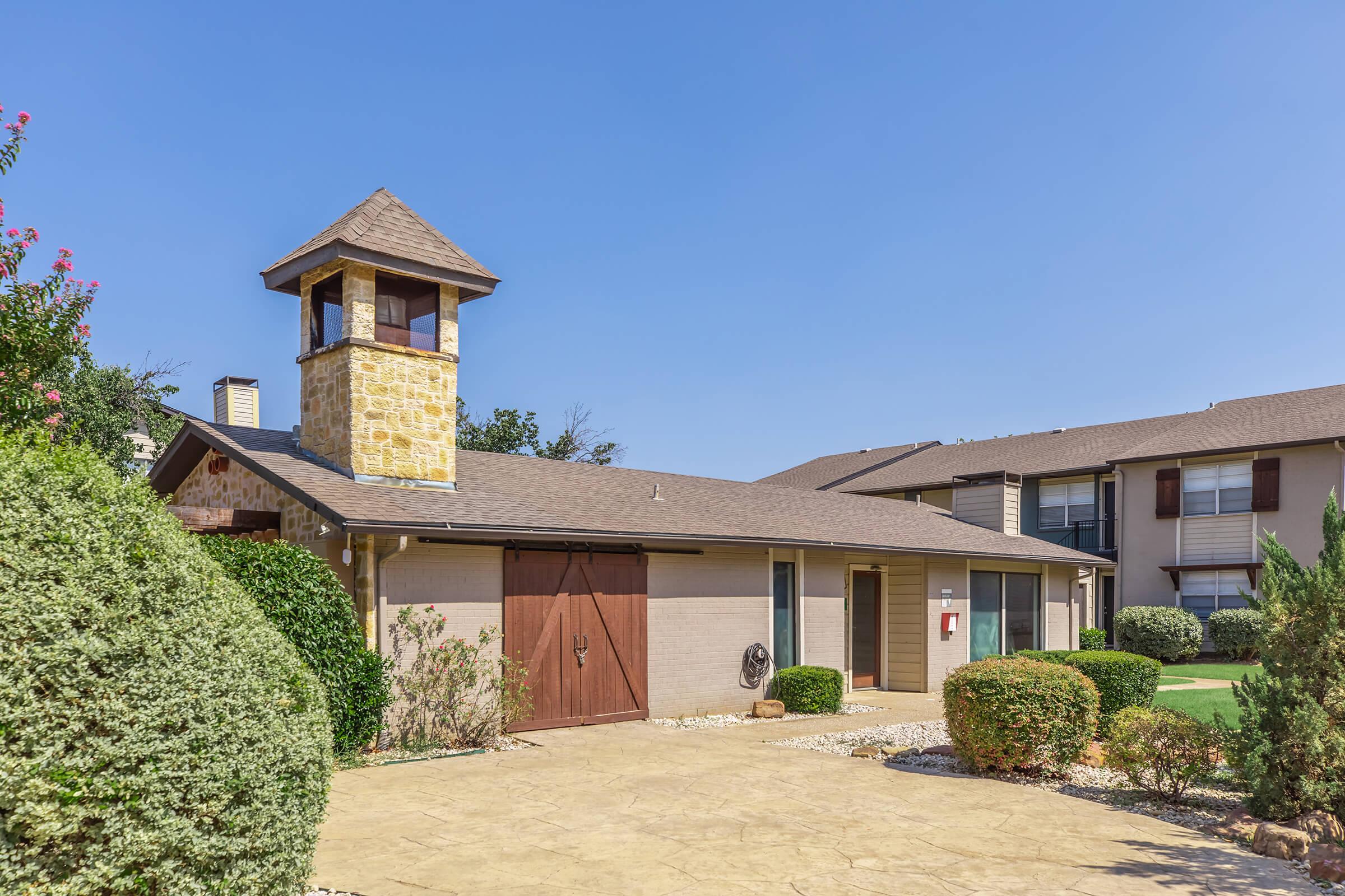 A quaint, single-story building with a brown exterior and a stone tower feature. Surrounding the building are well-maintained shrubs and landscaping. The sky is clear and blue, indicating a sunny day.