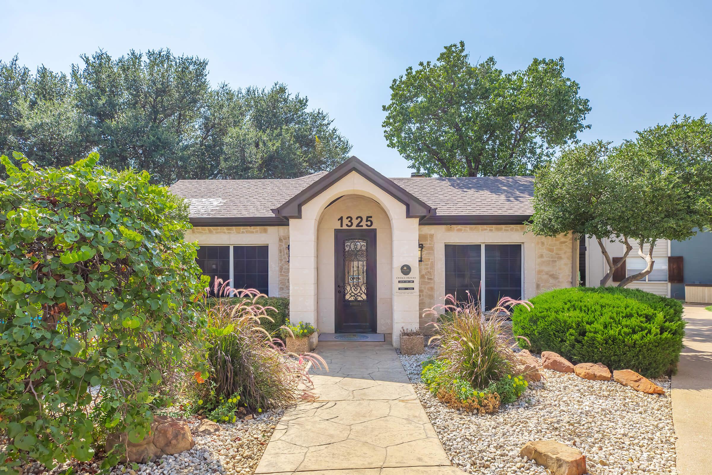A charming single-story house with a light stone facade, a decorative front door, and large windows, surrounded by lush green landscaping, including bushes and ornamental grasses. The pathway to the front door is paved with stones, leading to a welcoming entryway.