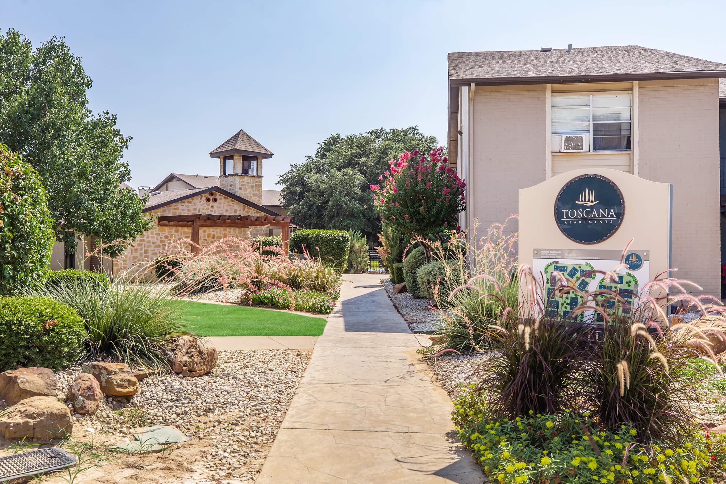 Pathway leading to Toscana Apartment Homes, featuring landscaped gardens with ornamental grasses and flowers. A stone pavilion is visible in the background, alongside buildings with a warm exterior. The community sign is prominently displayed at the forefront. Clear blue sky enhances the inviting atmosphere.
