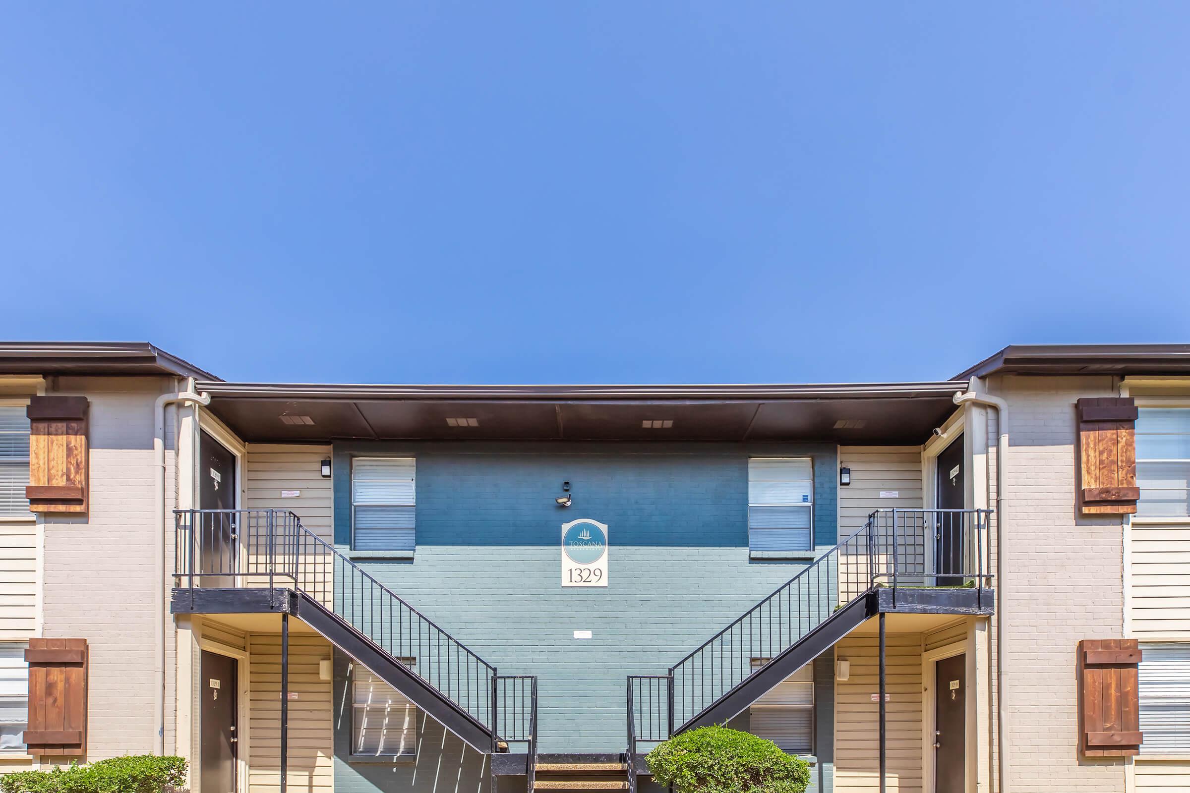 Two-story apartment building with a blue exterior and white trim. The building features two sets of stairs leading to an upper level, with metal railings. Each doorway is adorned with brown shutters. A clear blue sky serves as the backdrop. A sign above the middle door displays the number "1329."