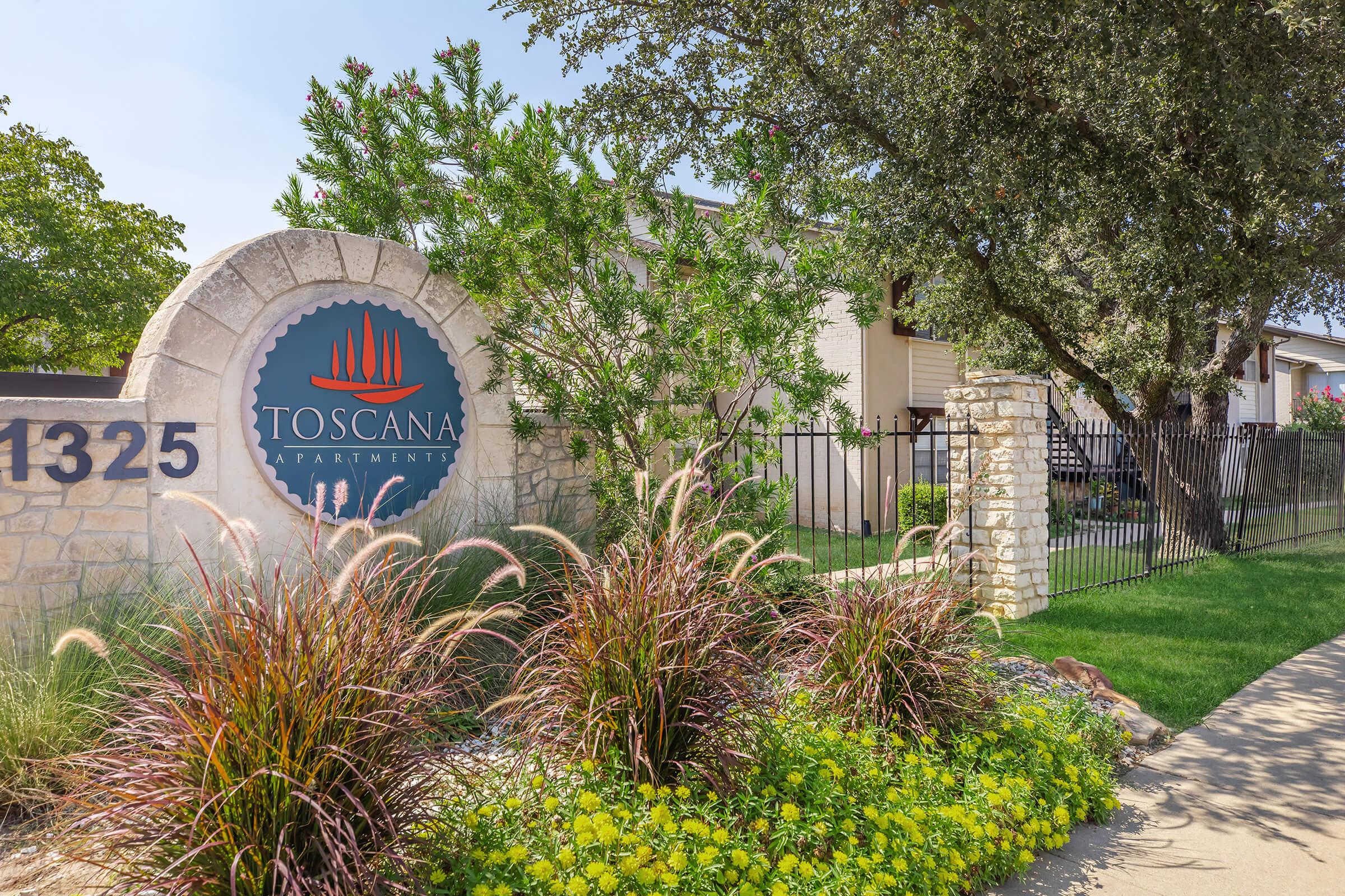 Sign displaying "Toscana Apartments" in front of the apartment complex, featuring a stone archway and lush landscaping with ornamental grasses and vibrant green plants. The scene is sunny, highlighting the entrance path and decorative elements around the sign.