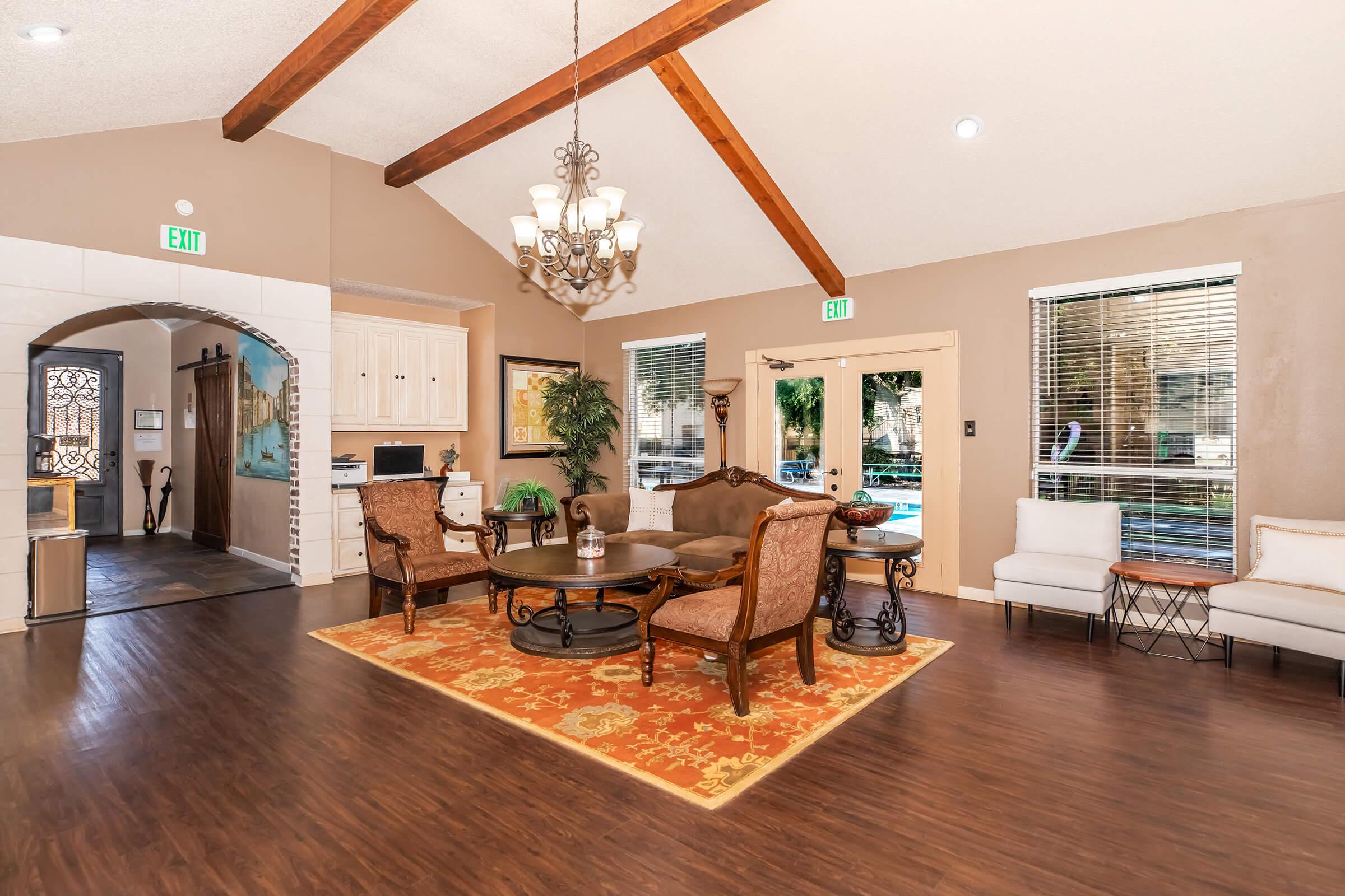 A spacious and elegantly designed living area featuring a high ceiling with exposed wooden beams. Furnished with a large area rug, comfortable seating, and decorative lighting, the room has large windows allowing natural light and a view of the outdoors. The decor is warm and inviting, suitable for relaxation.
