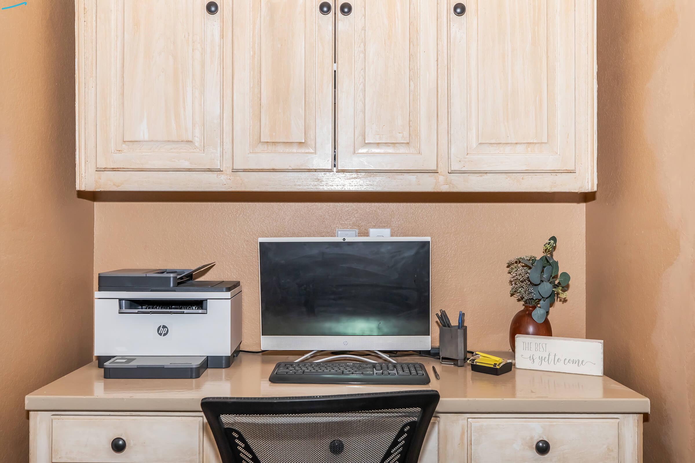 A home office desk setup featuring a desktop computer monitor, a printer, a small potted plant, and various stationery items. The desk is light-colored with a simple design, and there are wooden cabinets above it. The wall behind is painted a warm, muted color.
