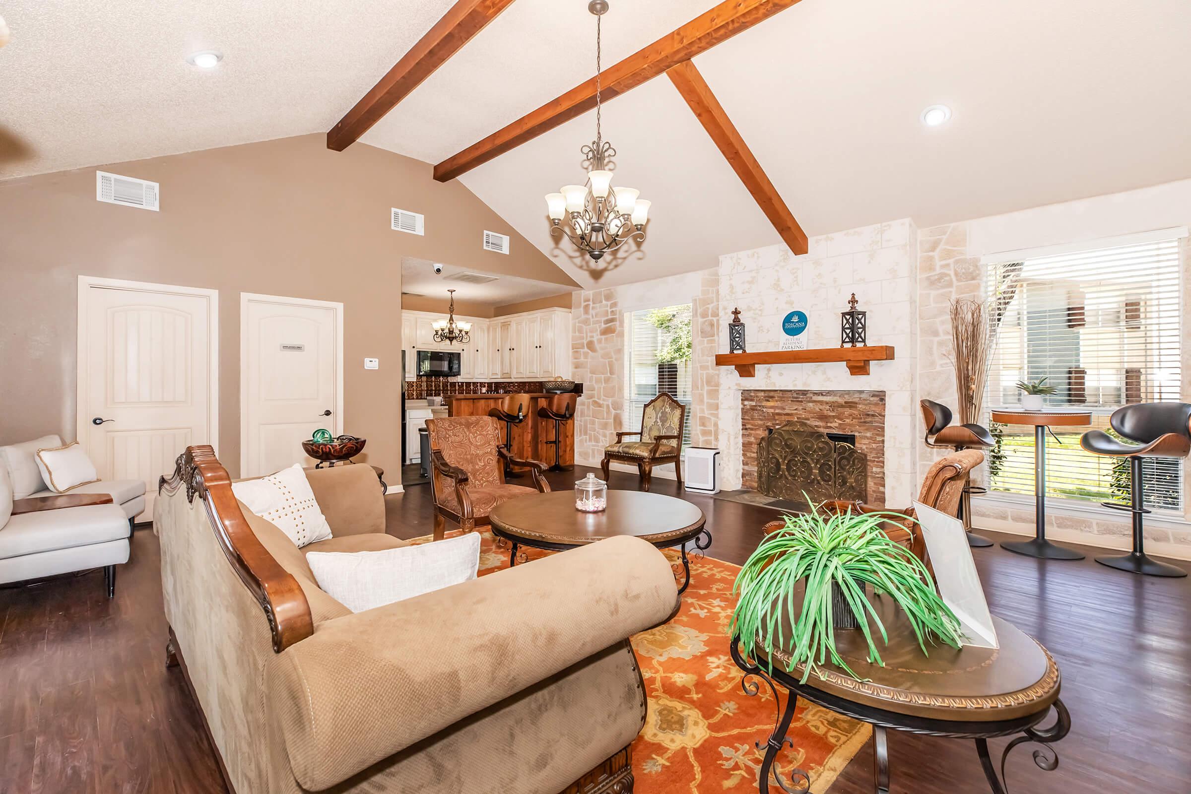A spacious living room featuring vaulted ceilings with wooden beams, a cozy fireplace, comfortable seating, and a decorative rug. Natural light flows in through large windows, highlighting the warm color palette and elegant decor. A dining area is visible in the background.