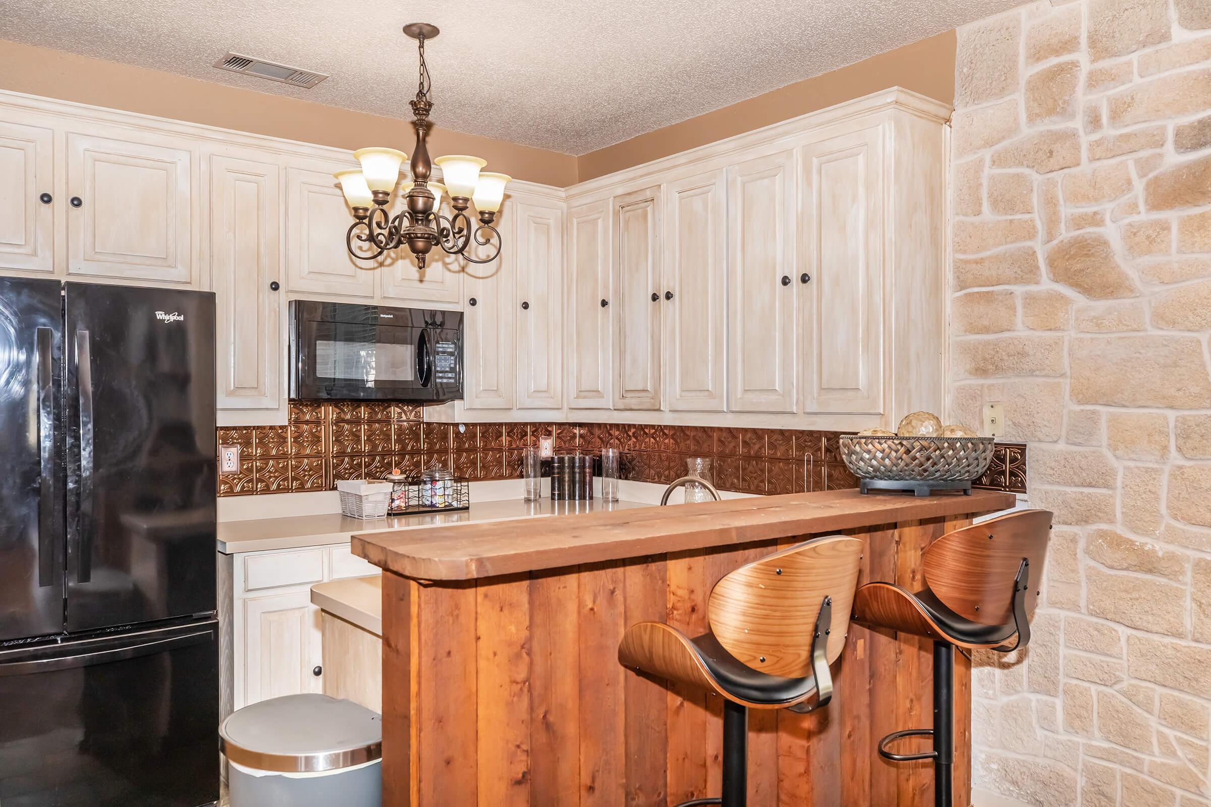 A modern kitchen featuring white cabinetry with a rustic finish, a black refrigerator, and a bar area with two wooden stools. The countertop is made of natural wood, and there's a decorative bowl on the surface. The back wall has a textured stone appearance, creating an inviting atmosphere.