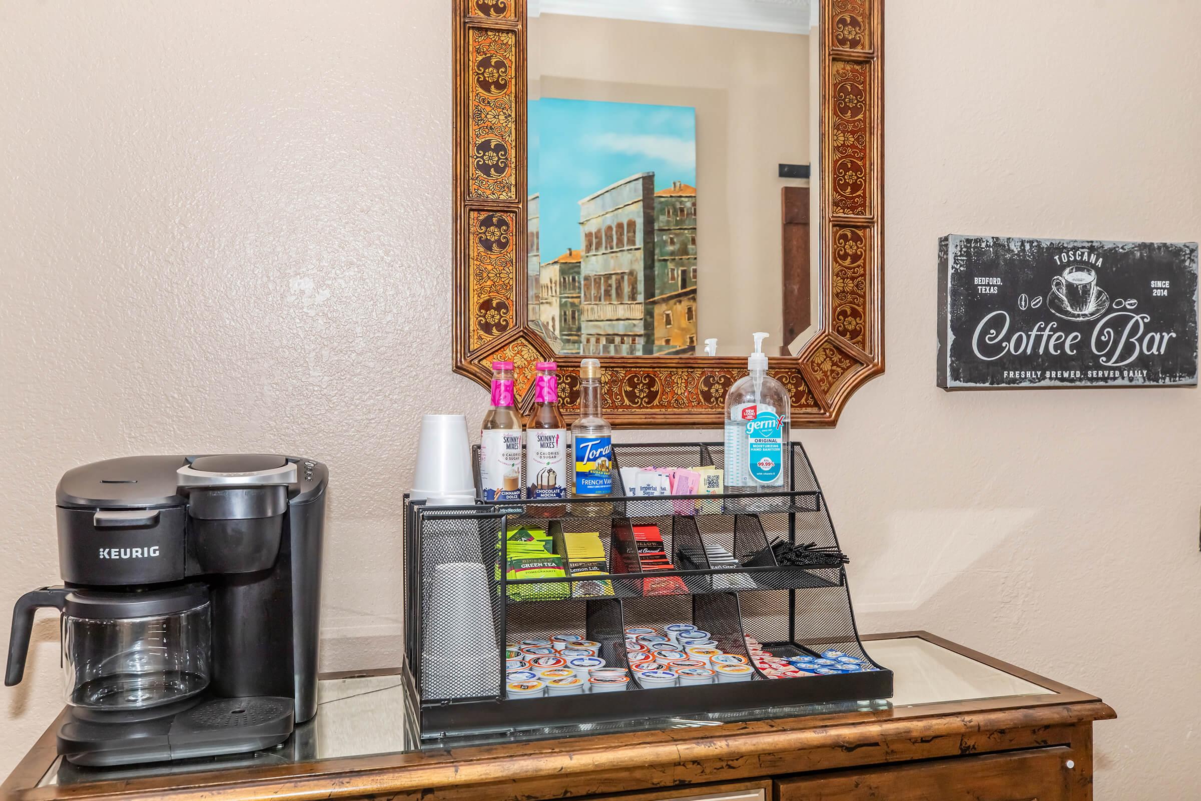A coffee station featuring a Keurig coffee maker, a decorative mirror, and a display rack with various coffee supplies, including cups, sugar packets, and creamers. Hand sanitizer is also available on the counter, alongside a "Coffee Bar" sign. The background includes a painting of a building.
