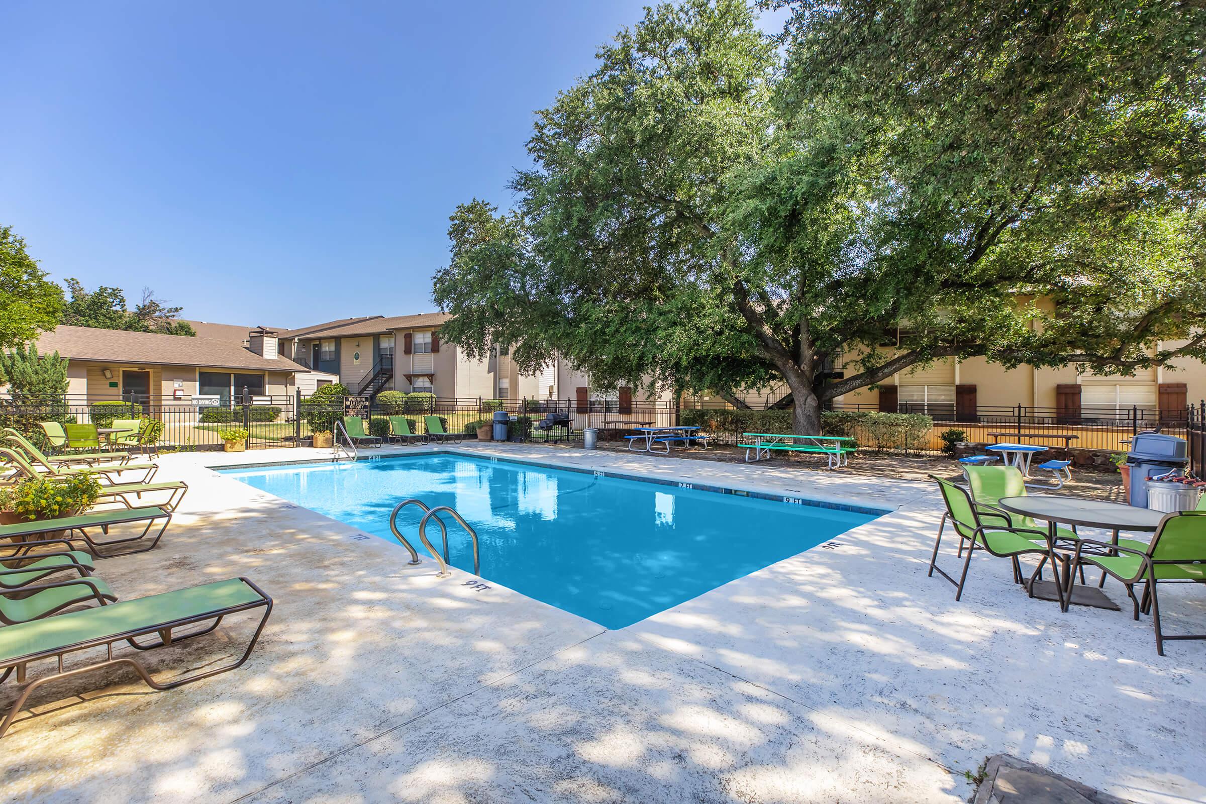 A sunny outdoor pool area surrounded by greenery, featuring clear blue water, lounge chairs, and tables. The pool is bordered by a concrete deck, with a few trees providing shade. Nearby buildings can be seen in the background, creating a welcoming atmosphere for relaxation.