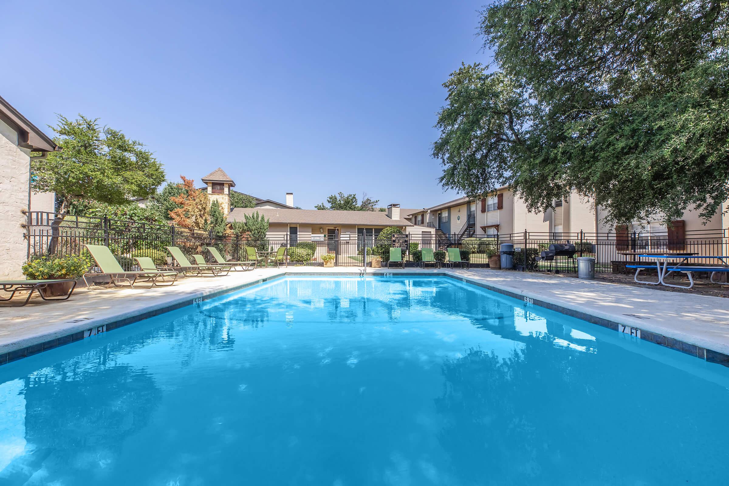 A clear blue swimming pool surrounded by lounge chairs and greenery. The pool area includes a fence, with a few buildings visible in the background. The sky is clear and blue, creating a bright and inviting ambiance.