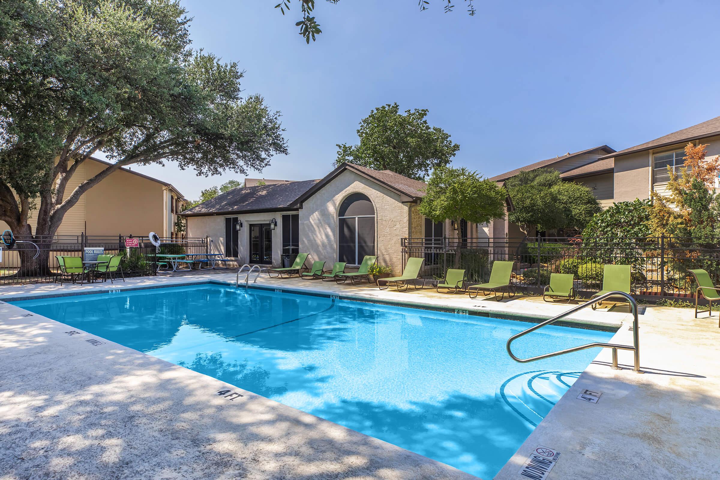 A bright outdoor swimming pool surrounded by green lounge chairs and shaded by trees. The pool features a staircase entry and is bordered by grass and a fence, with a residential building visible in the background against a clear blue sky.