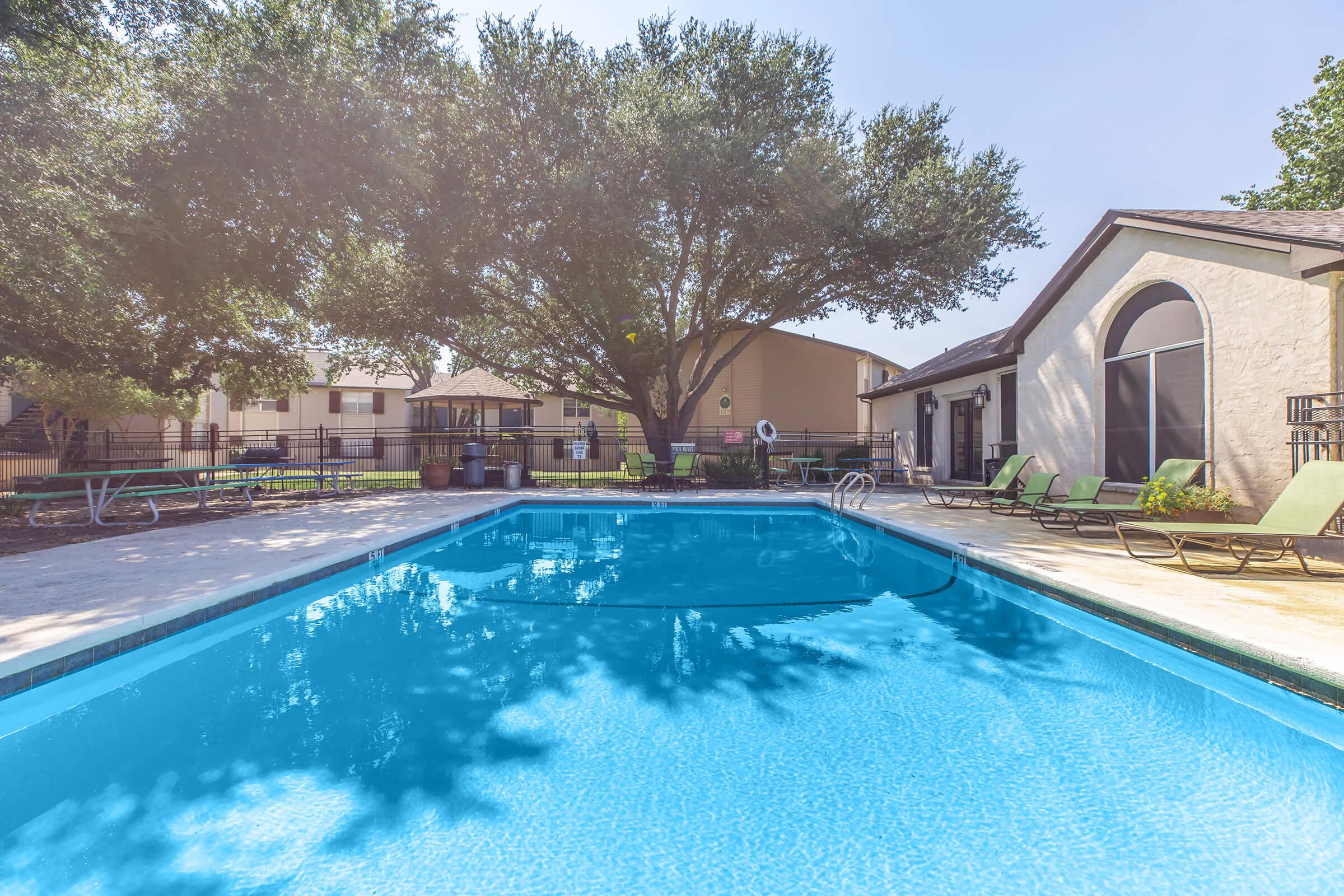 A clear blue swimming pool surrounded by a spacious patio, with lounge chairs arranged around it. In the background, there are trees and residential buildings, creating a peaceful outdoor atmosphere. The sky is bright and sunny, enhancing the inviting scene of leisure.