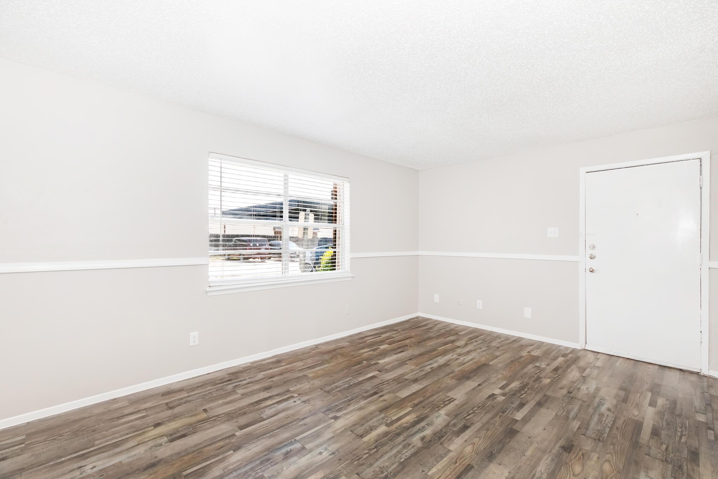 A spacious, empty room featuring light-colored walls and a textured ceiling. Natural light enters through a window with white blinds, and a door is visible on one wall. The flooring is made of light wood, creating a warm atmosphere. The space is clean and ready for furnishings.