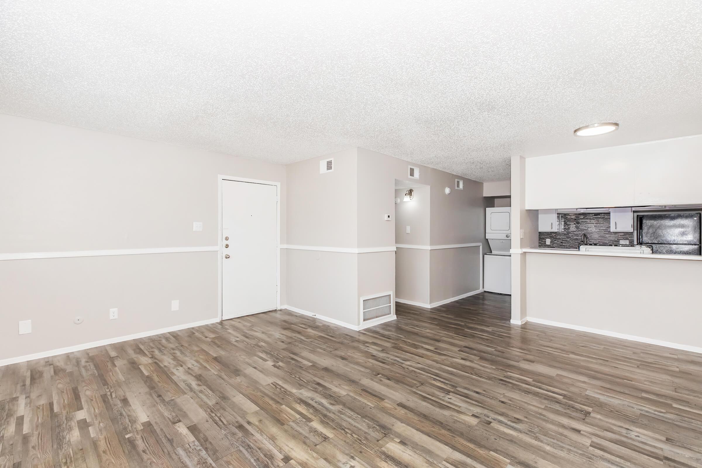 A spacious, empty living area with light-colored walls and a textured ceiling. The floor features modern hardwood-style flooring. There is a door leading outside, and an open archway connects to a kitchen area in the background. Natural light brightens the room, creating a welcoming atmosphere.