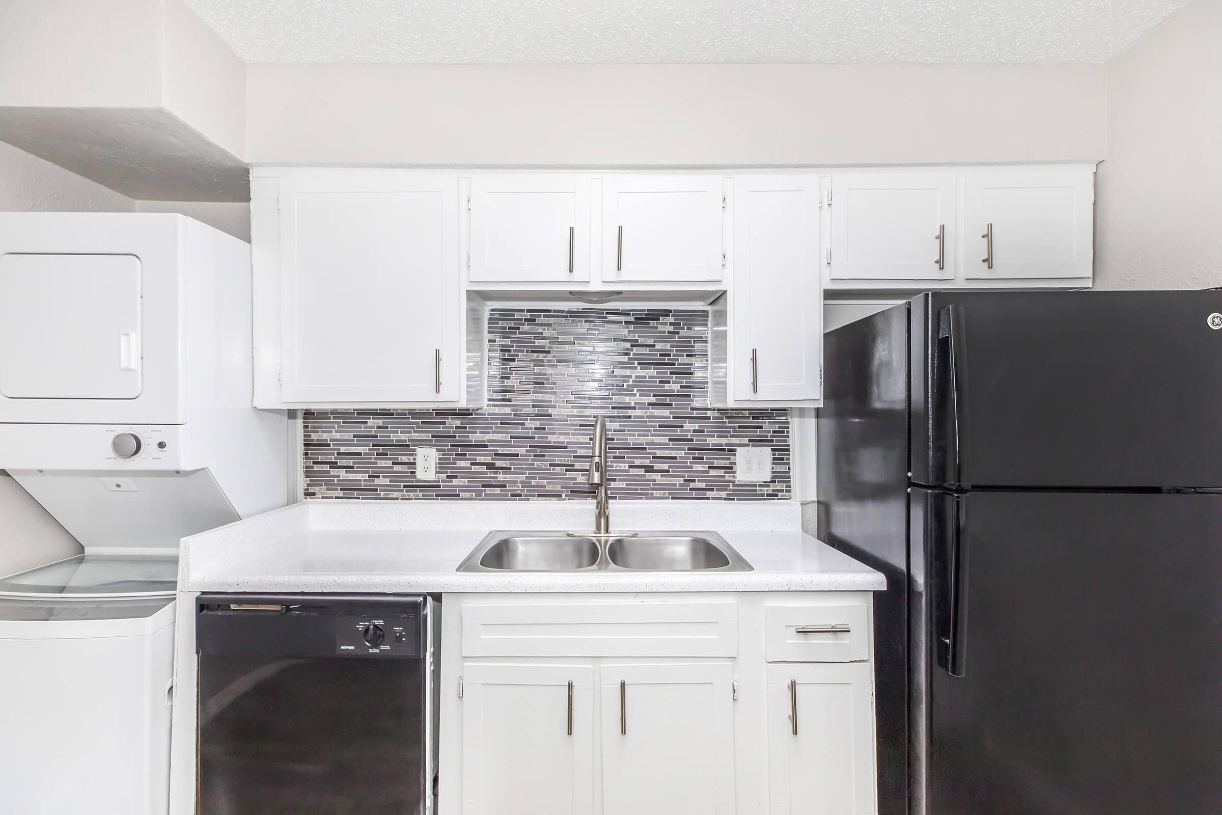A modern kitchen featuring white cabinetry, a double sink, and stainless steel appliances. The countertop is white, and there's a black refrigerator and dishwasher. A gray and white tiled backsplash adds a contemporary touch. A washer and dryer are visible in the corner, enhancing the utility of the space.