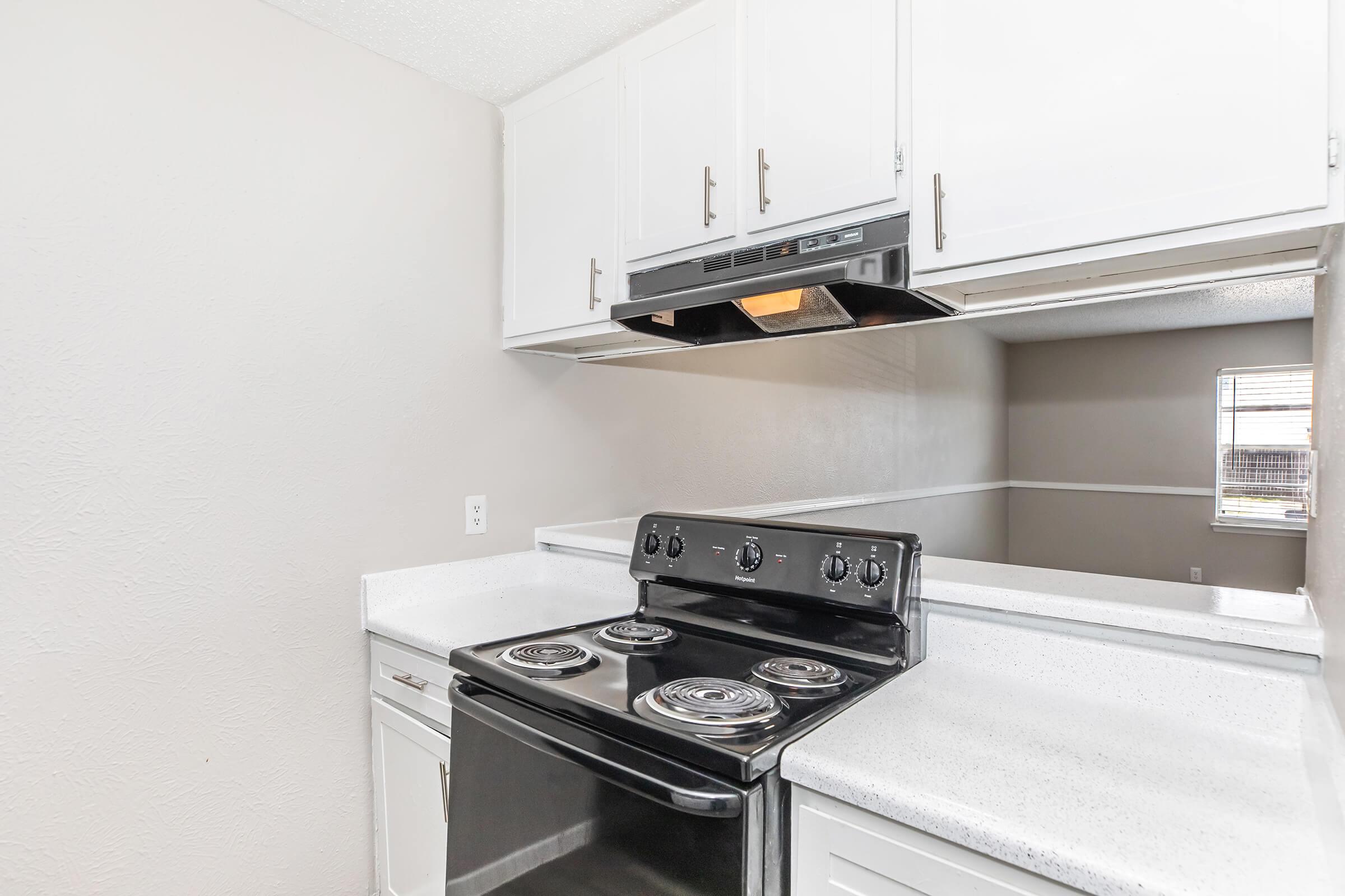 A kitchen featuring a black electric stove with four burners, a microwave above, and white cabinetry. The countertops are light-colored, and the walls are painted a soft grey. Natural light is visible from a nearby window, enhancing the clean and modern aesthetic of the space.