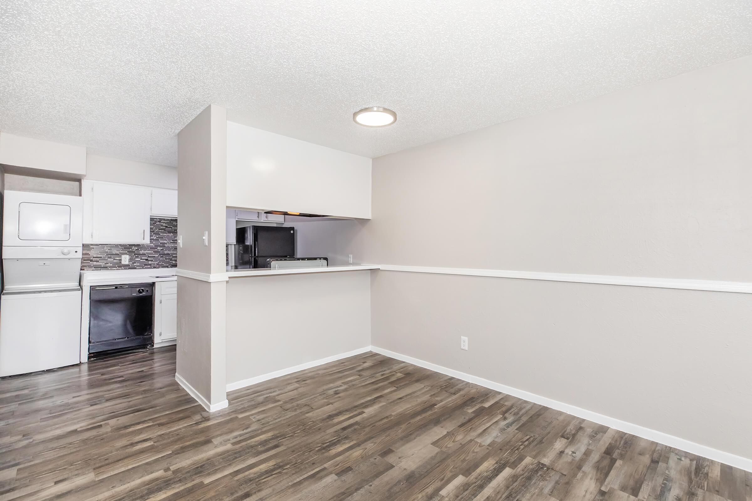 A spacious, open-concept living area with light-colored walls and modern laminate flooring. To the left, there is a laundry area with a washer and dryer, while a kitchen with dark appliances is partially visible in the background. The overall atmosphere is bright and inviting.
