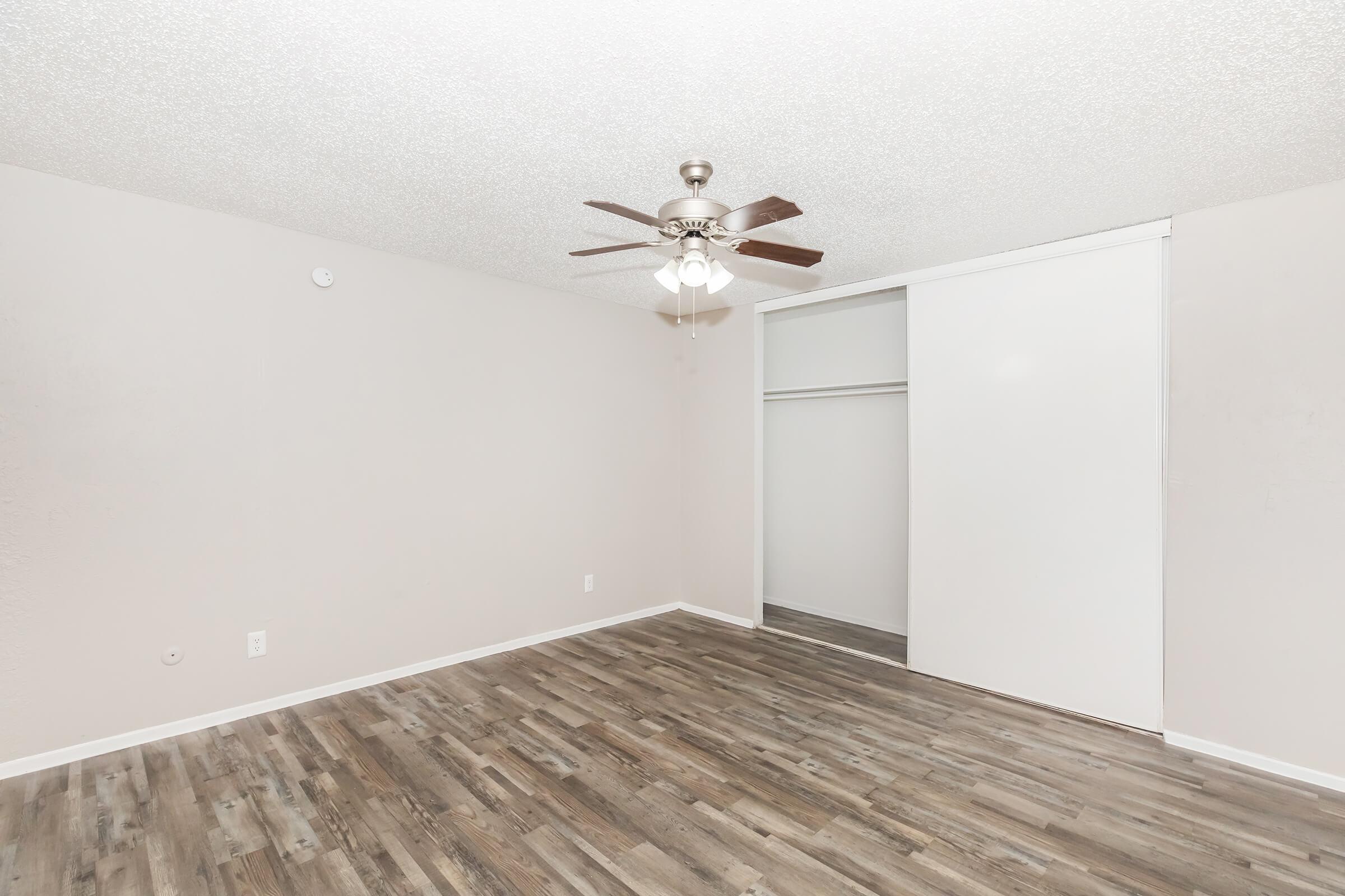 A spacious, empty room featuring light-colored walls, a ceiling fan, and a wooden floor. There is a large white closet with sliding doors on one side, providing ample storage space. The overall atmosphere is bright and airy, suitable for various uses.