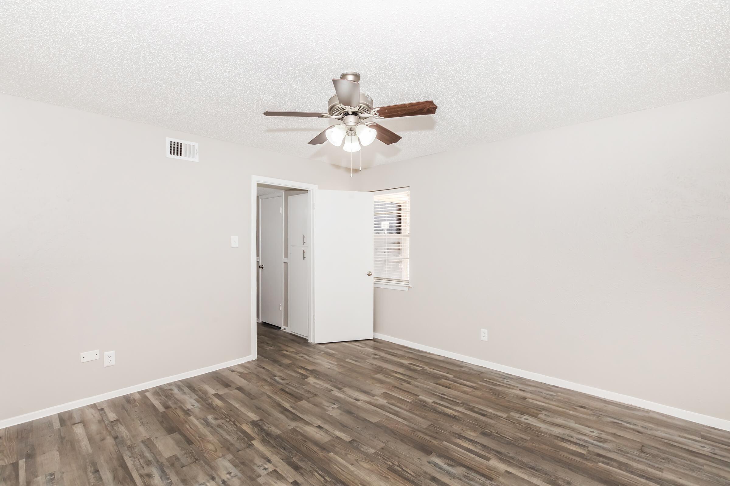 Empty room featuring a light brown wood floor, pale beige walls, and a ceiling fan with three lights. There's a white door partially open, leading to another space, and a window allowing natural light in. The overall ambiance is bright and airy.