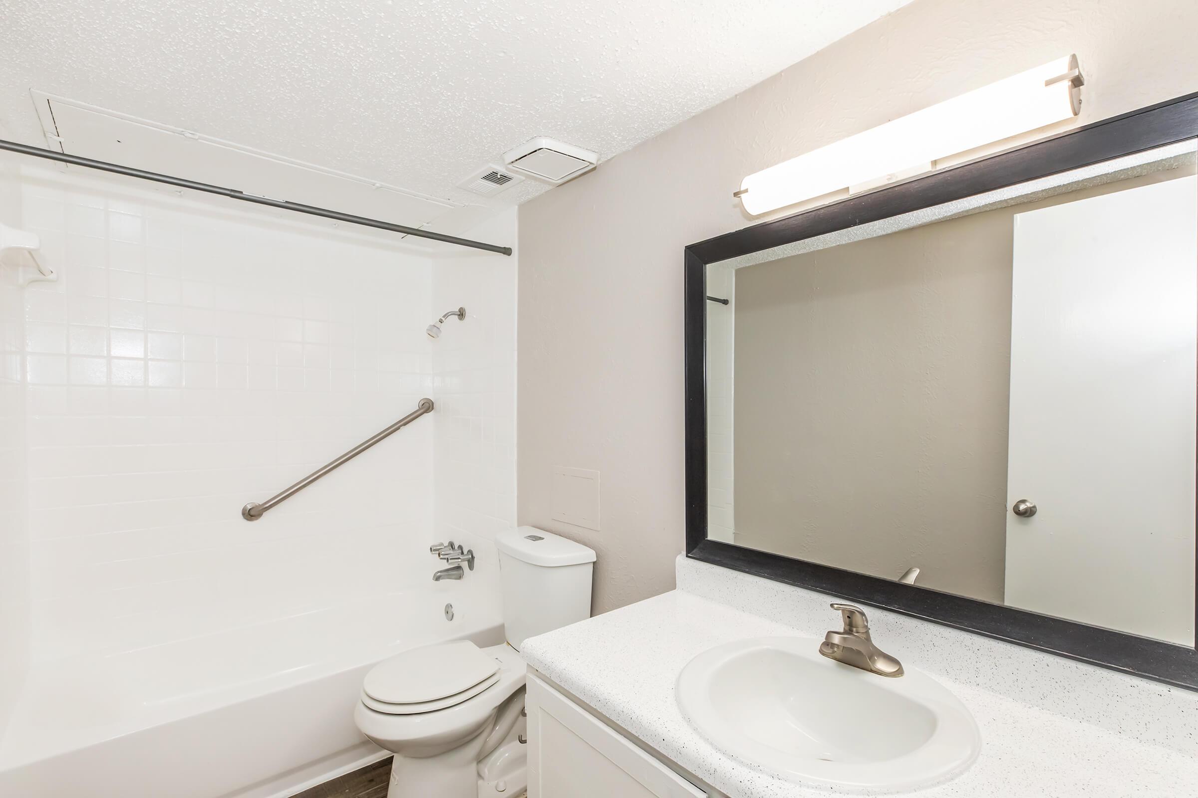 A clean, modern bathroom featuring a white bathtub with a showerhead, a toilet, a sink with a brushed metal faucet, and a large mirror above the sink. Soft lighting from a mounted fixture illuminates the space, which has light-colored walls and a simple design.