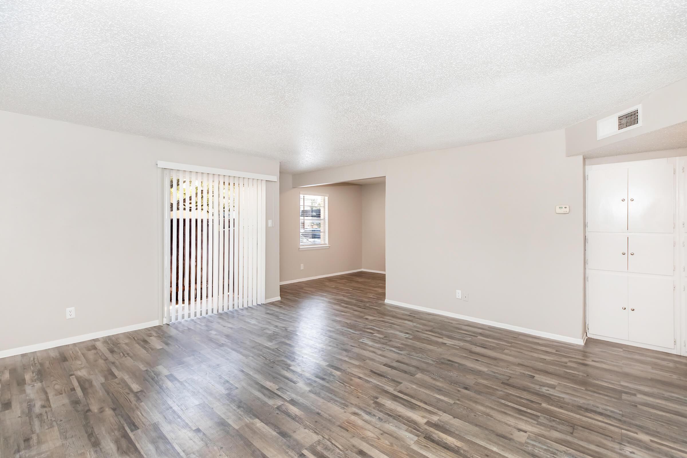 Spacious interior of a light-colored living area featuring laminate flooring. One wall has vertical blinds leading to an outdoor space, while another wall includes a small doorway to an adjoining room. White walls enhance the brightness, creating an open and inviting atmosphere.