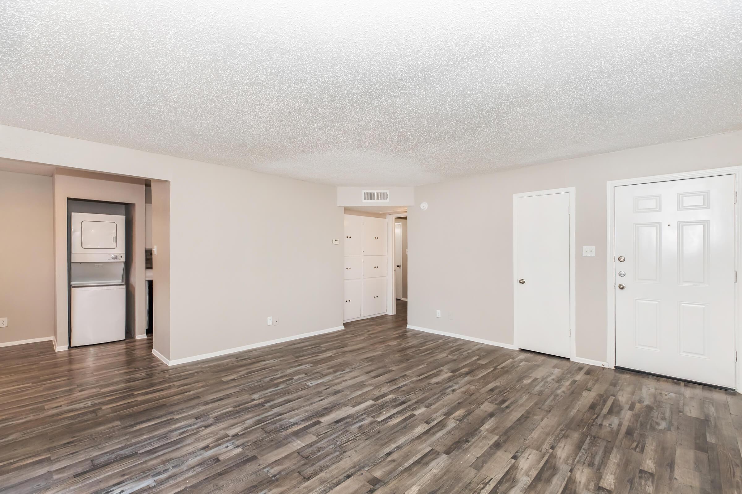 A spacious, light-colored living room featuring hardwood-style flooring. There is a door leading to an entrance, a closet, and a laundry area visible in the background. The walls are painted a soft neutral tone, contributing to a bright and airy atmosphere.