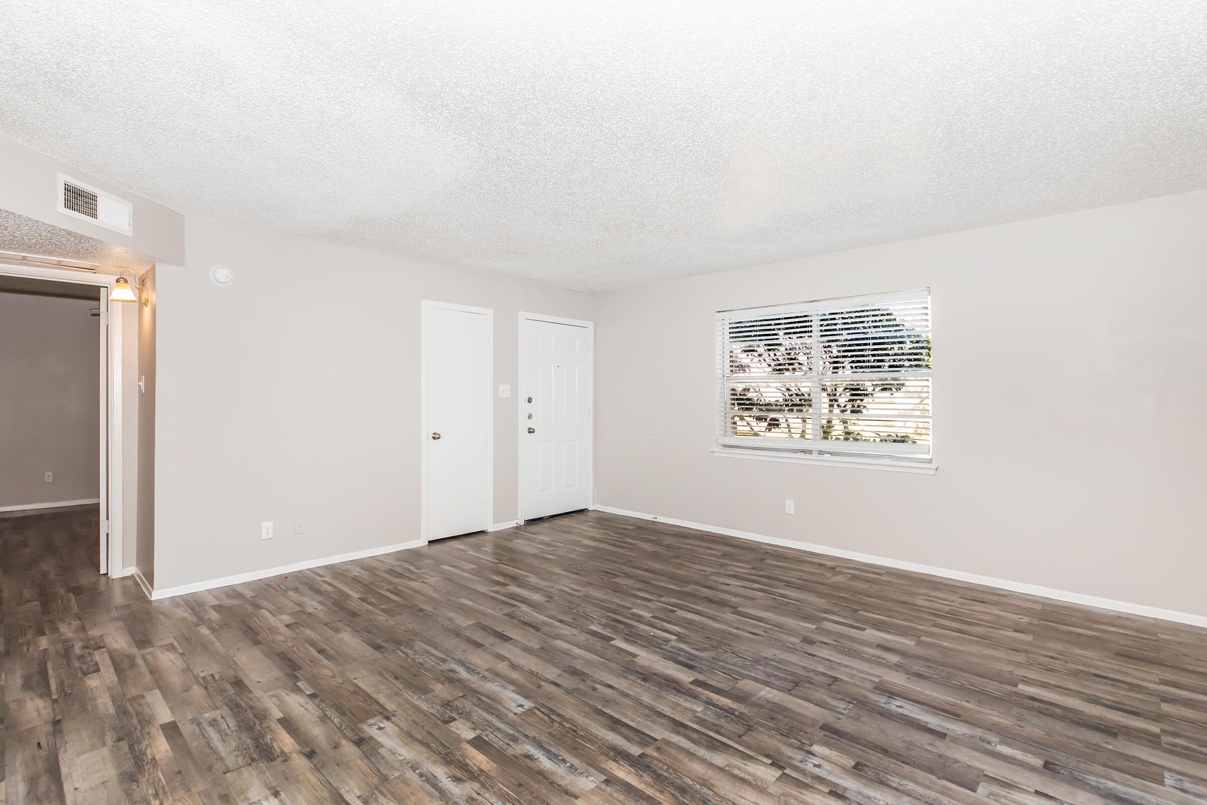 Spacious, empty living room featuring light-colored walls, laminate flooring, and a large window with blinds. A door leads to the entrance, and another door provides access to an adjacent room. The room is well-lit and designed for various furniture arrangements.