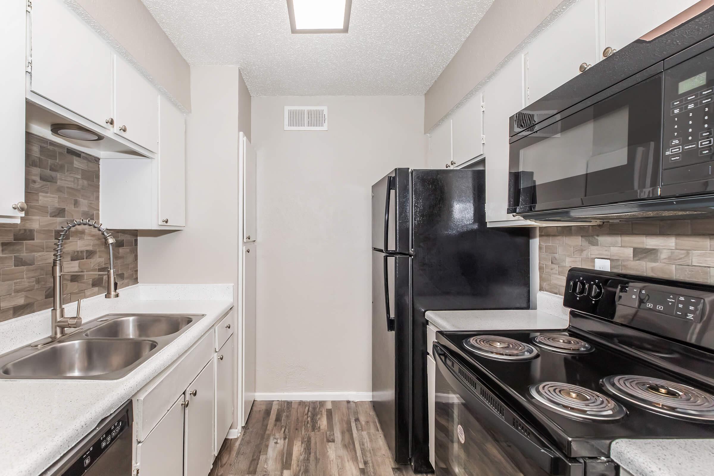 Kitchen with white cabinets and a black refrigerator, microwave, and stove. The countertops are light-colored, and there are dual sinks. The backsplash features a tiled design. The walls are painted in a neutral tone, and the flooring has a wood-like appearance. A door leads to another area of the home.