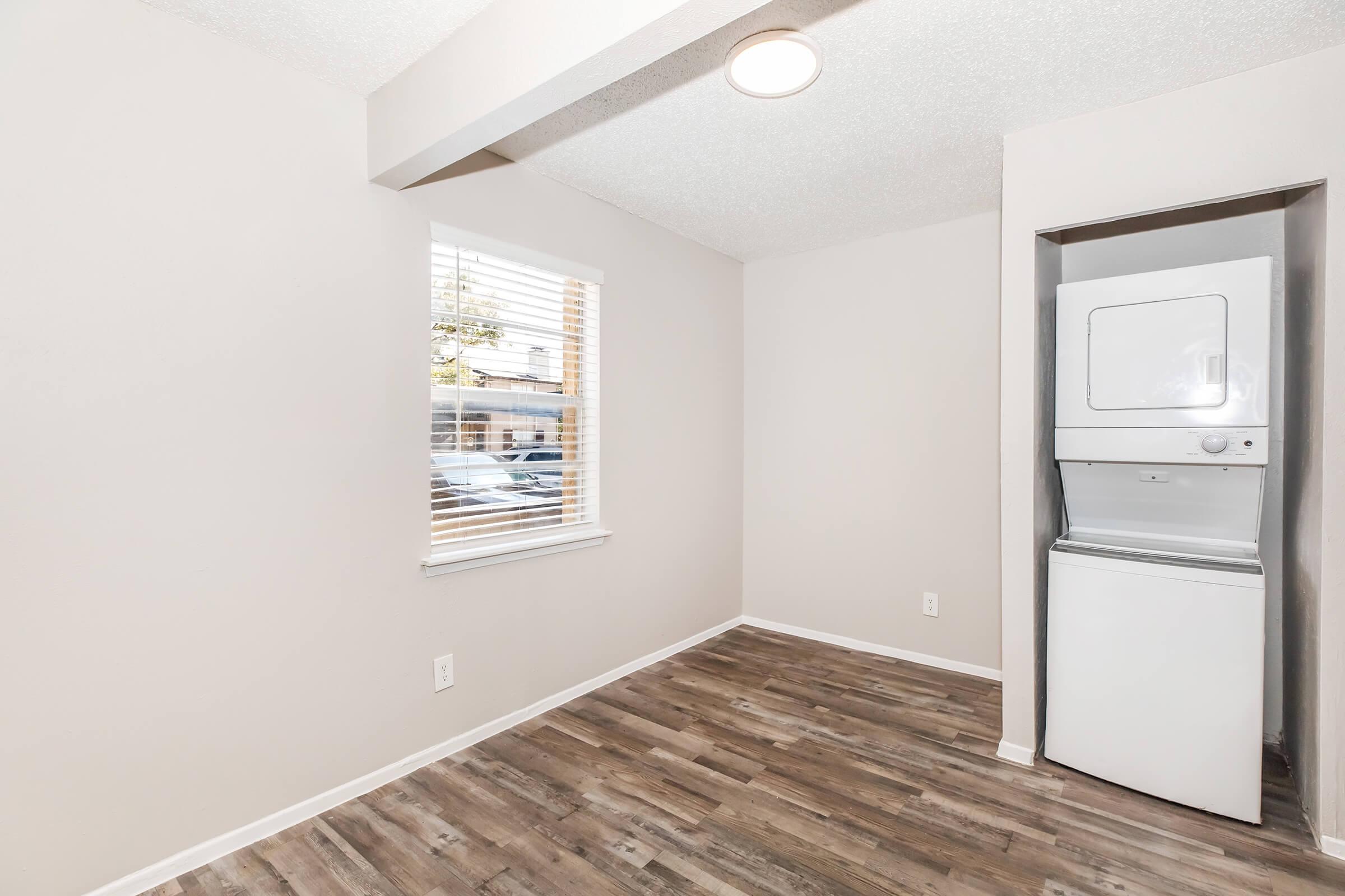A small, light-colored room with a window featuring blinds, revealing a view of the outdoors. The floor is covered with laminate flooring, and a stacked washer and dryer unit is located in the corner. The walls are painted in a neutral shade, creating a bright and airy atmosphere.