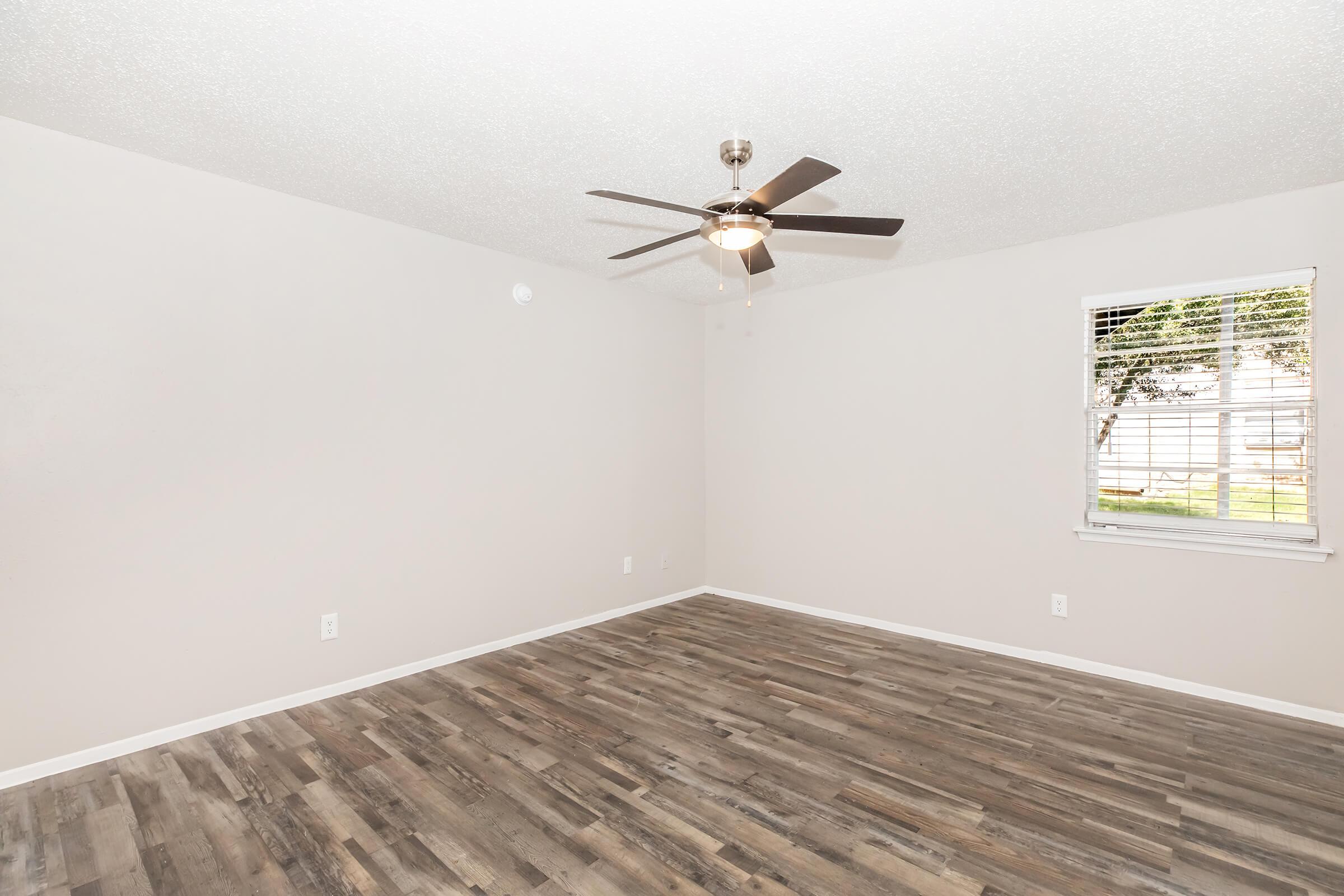 A spacious, empty room with light-colored walls and a textured ceiling. The flooring features wood-like planks, and a ceiling fan is installed in the center. A window with blinds allows natural light to enter, offering a view of greenery outside. The room is open and bright, ready for furniture.