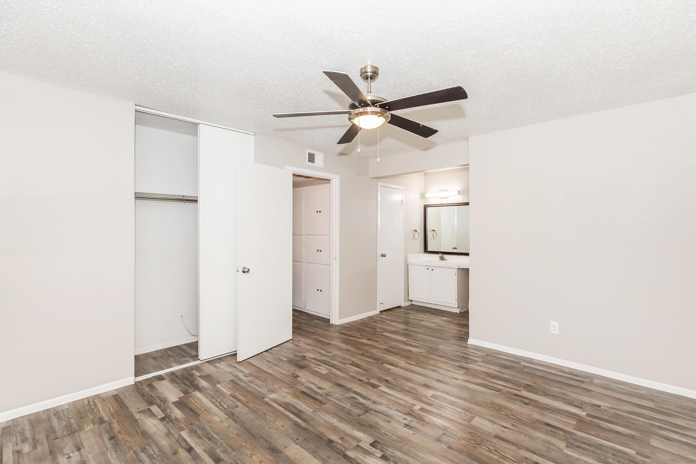 A spacious, light-filled room featuring a ceiling fan, laminate flooring, and neutral-colored walls. The room includes a closet with double doors on the left, and a separate area with a bathroom vanity mirror and cabinets. The overall decor conveys a clean and contemporary design.