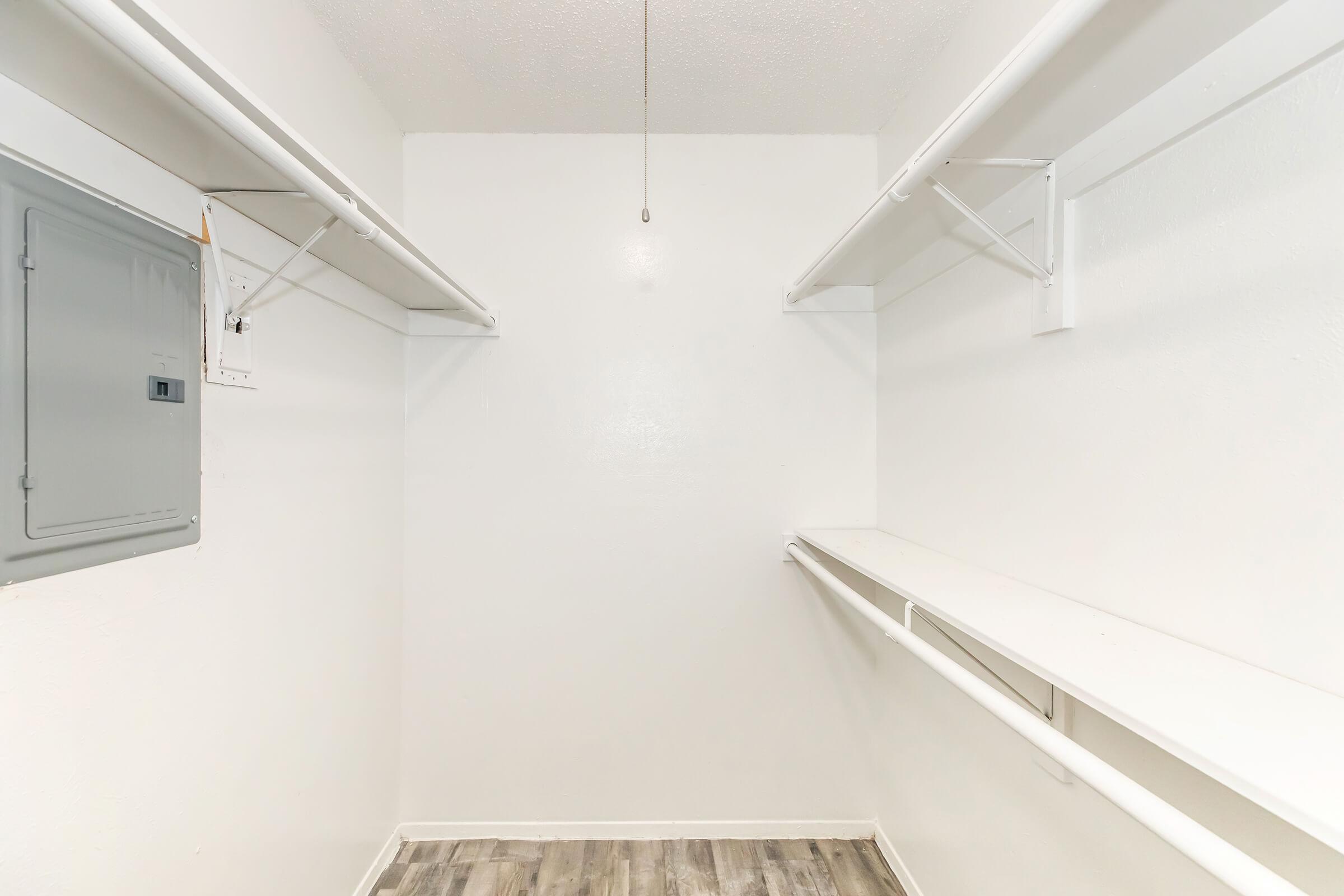 A clean, empty closet with white walls, featuring two hanging rods on either side and a shelf along the back wall. A small electrical panel is visible on the left side. The floor is covered with light-colored, textured planks. The space is well-lit and uncluttered.