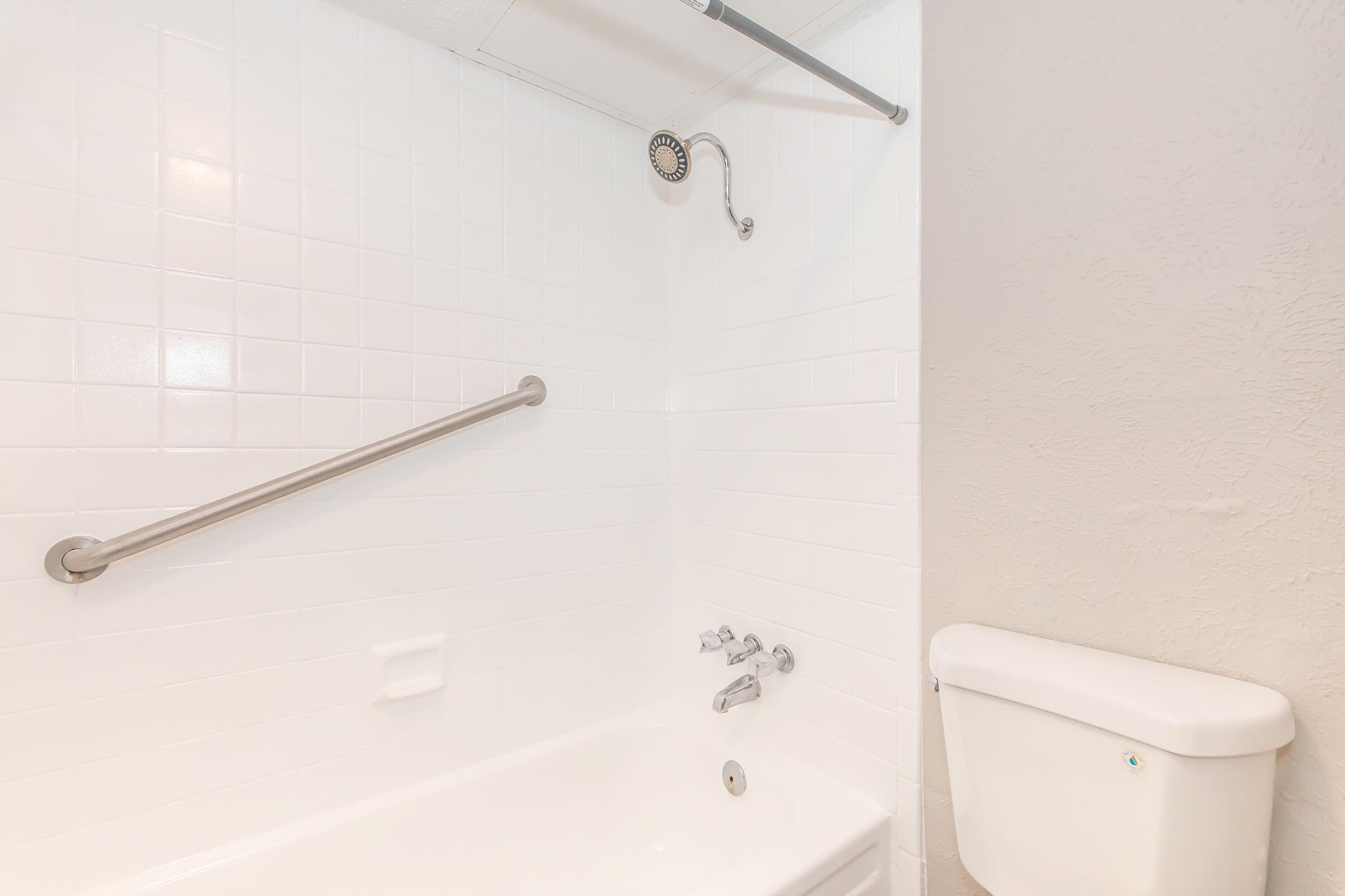 A clean, modern bathroom featuring a white tiled bathtub with a showerhead, grab bars for safety, and a white toilet. The walls are light-colored and the space appears well-lit and tidy, creating a serene atmosphere.
