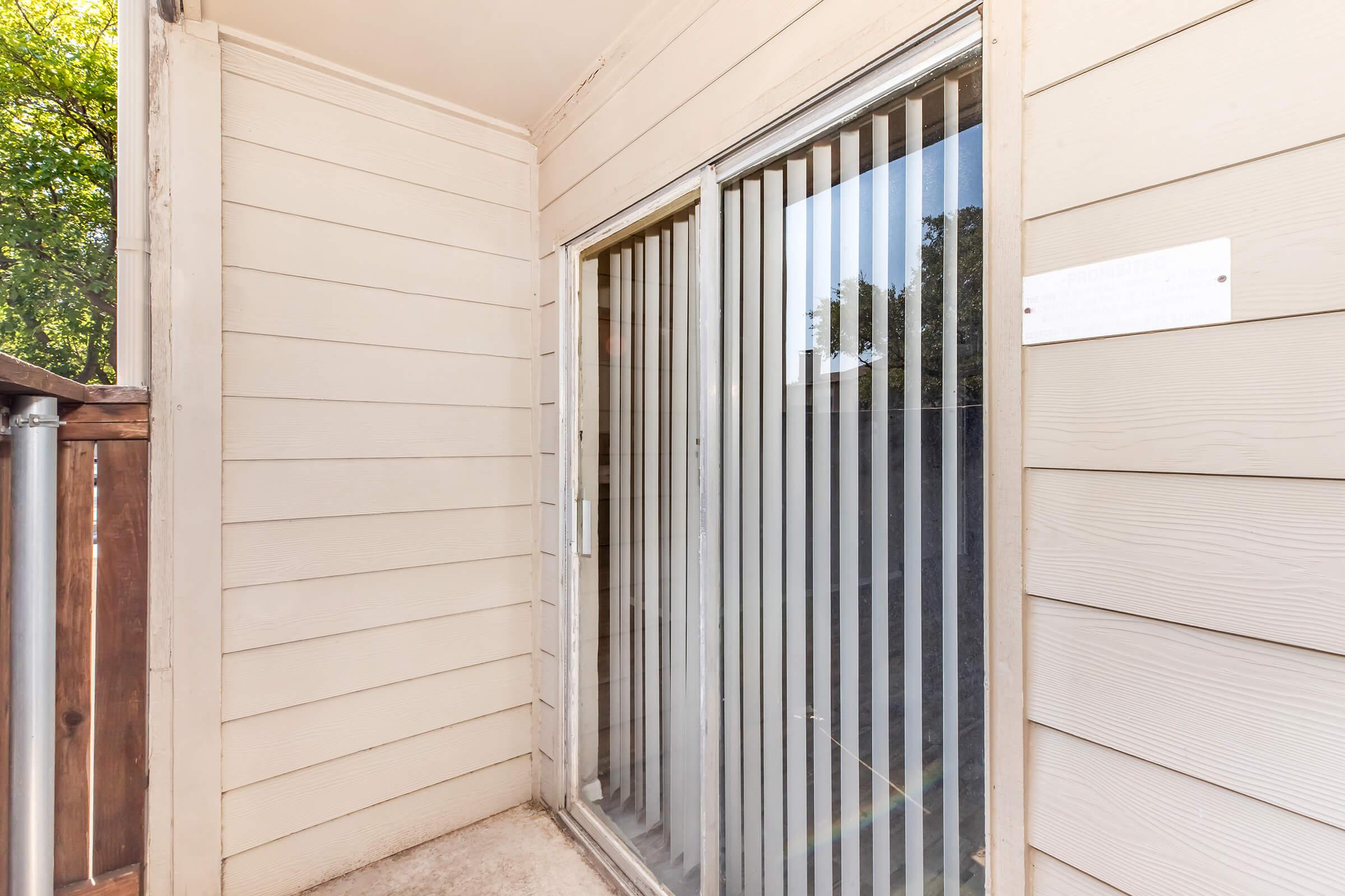 A corner of a balcony or patio featuring sliding glass doors with vertical blinds. The walls are light-colored wood or siding, and there is a small sign on the wall near the doors. Natural light filters in from outside, illuminating the space.