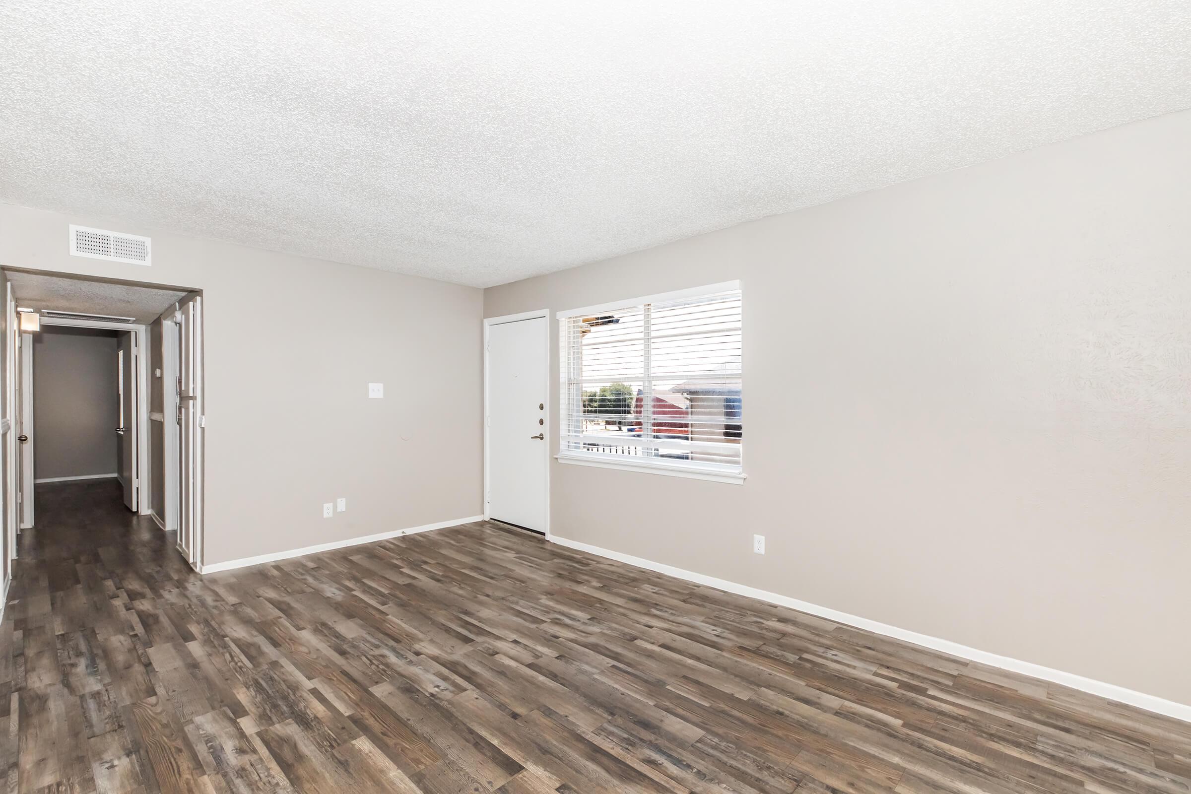 An empty room with light gray walls and a textured ceiling, featuring a door on the right leading outside, a window with blinds letting in natural light, and wooden flooring. A hallway entrance is visible on the left, suggesting additional adjoining rooms.