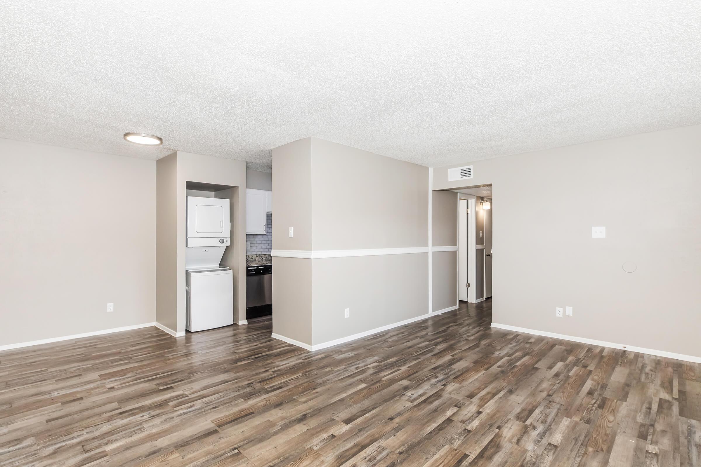 A spacious interior view of a living area with light gray walls and polished wooden flooring. In the background, there is a washer and dryer unit in a small nook, with a kitchenette visible to the left. The space is well-lit, featuring a ceiling light and a neutral color palette, creating a modern, open atmosphere.