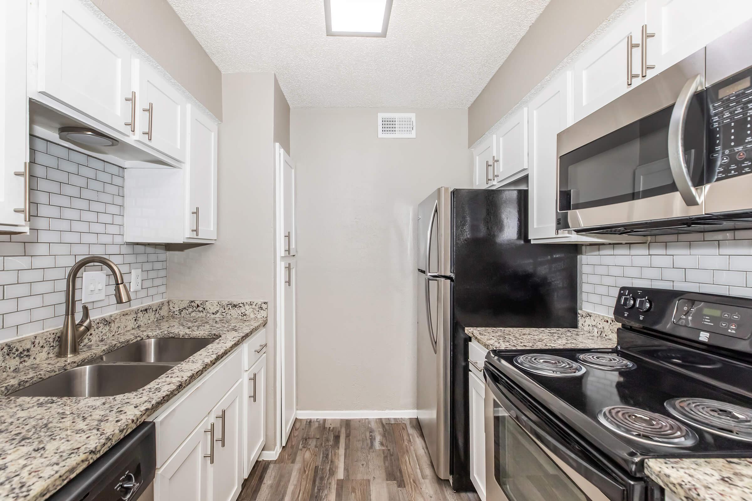 A modern kitchen featuring white cabinetry, granite countertops, stainless steel appliances, and a black refrigerator. The layout includes a double sink, an electric stove, and a microwave, with a decorative tile backsplash and wooden flooring. Natural light enters through a ceiling fixture.