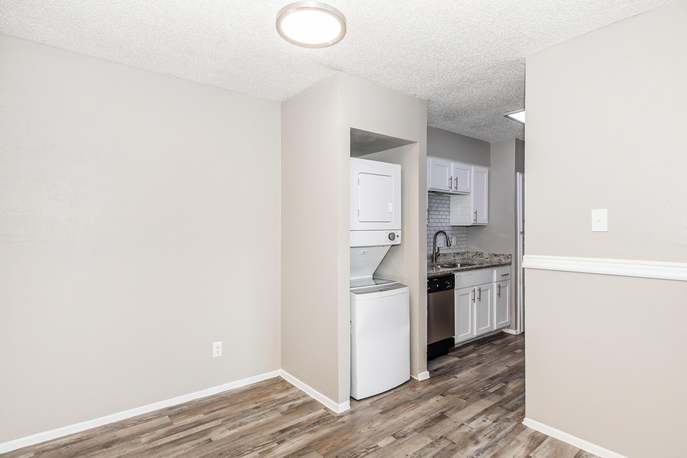 A modern kitchen area featuring gray walls, vinyl plank flooring, and a small laundry nook with a stacked washer and dryer. The kitchen includes white cabinets, a stainless steel sink, and a view into an adjacent room. Soft overhead lighting enhances the spacious feel.