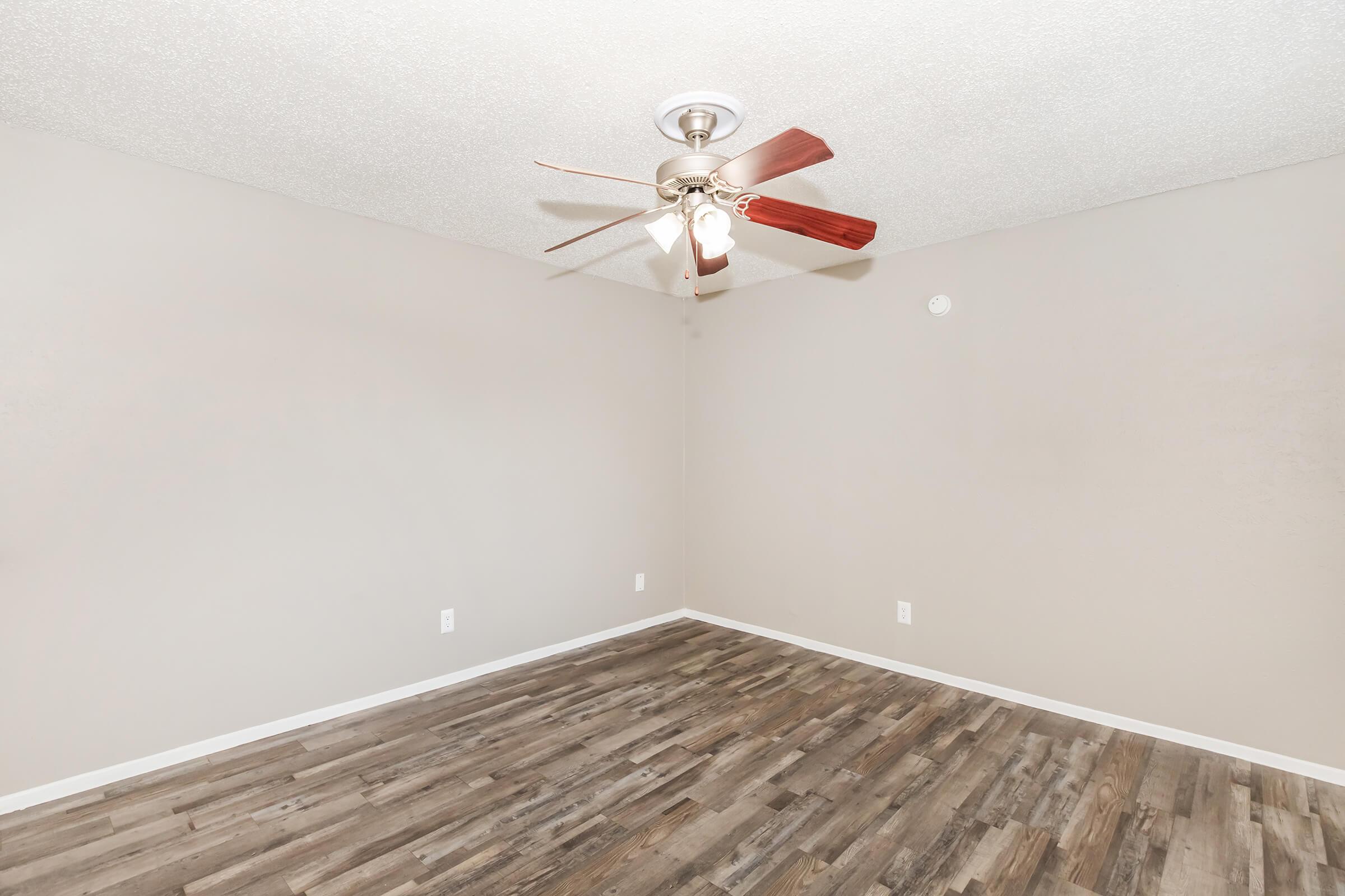 A spacious, empty room with light brown wooden flooring and a textured light gray wall. At the center, there is a ceiling fan with wooden blades and a light fixture. The room has a simple and clean aesthetic, suitable for various uses or decoration.