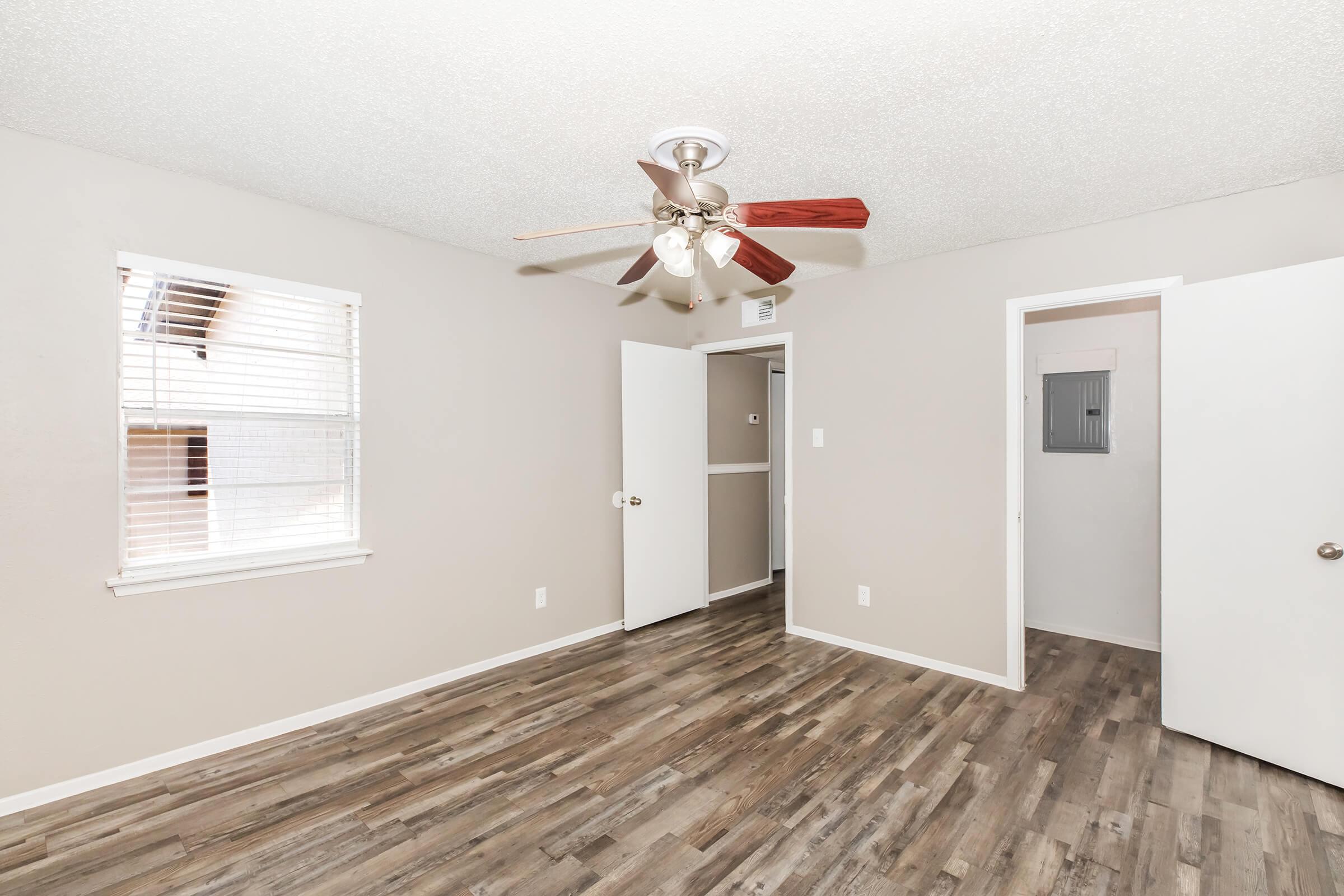 A well-lit room with light gray walls and wood laminate flooring. It features a ceiling fan with wooden blades, a window with blinds, and two open doors leading to other areas. The interior is simple and clean, creating a spacious feel.