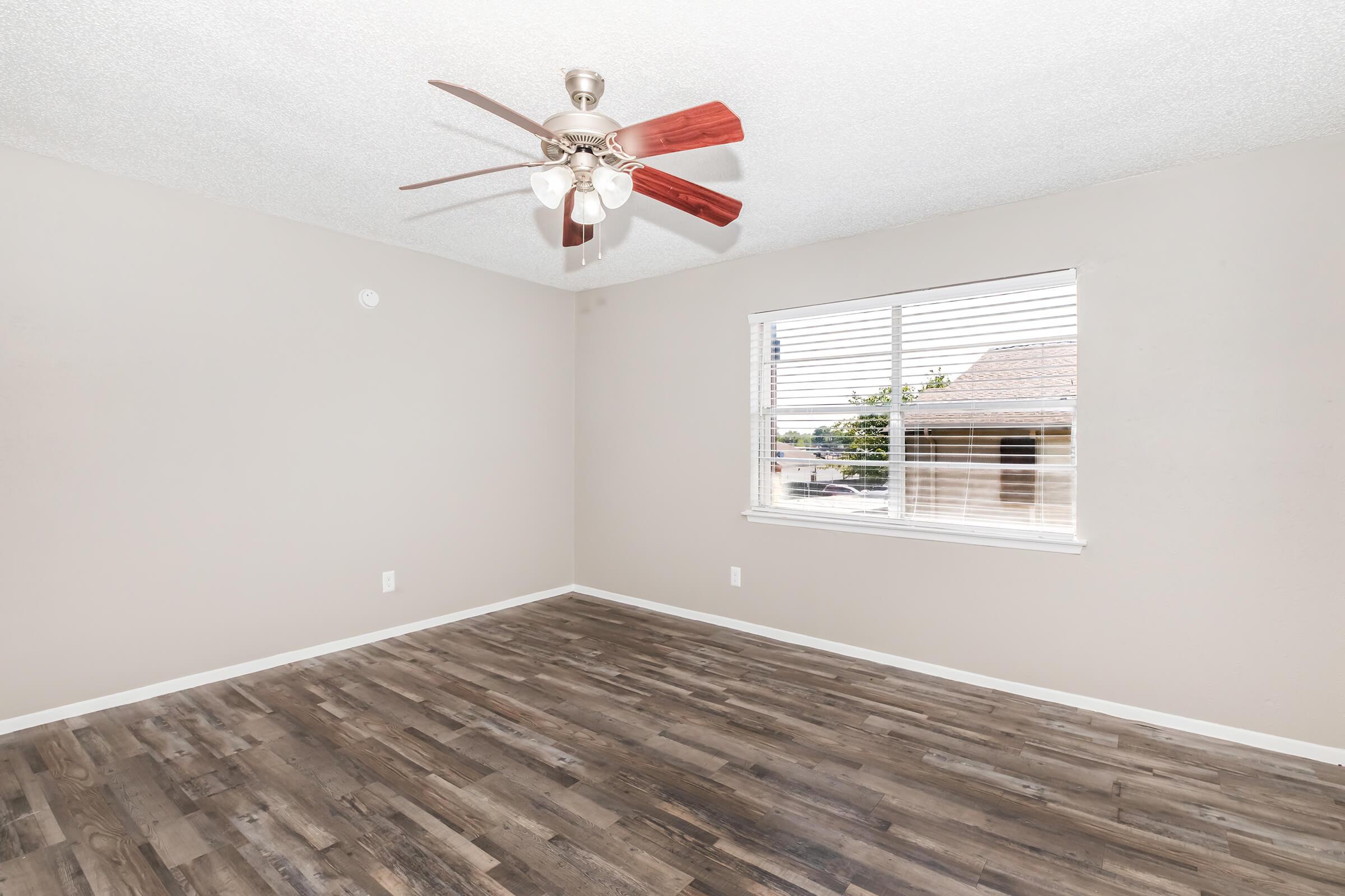 A well-lit, empty room featuring a ceiling fan with red blades. The walls are painted a light beige, and there is a window with blinds, providing a view outside. The floor is covered in dark laminate wood, creating a warm contrast with the lighter walls.