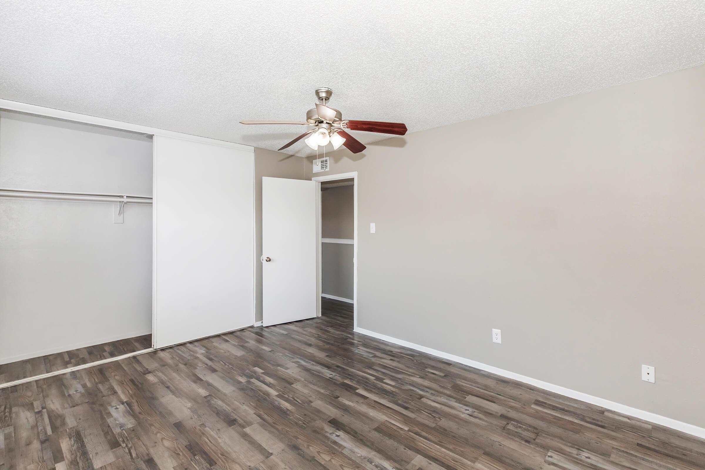 A room with light gray walls and wooden flooring. There's a ceiling fan with wooden blades. On one side, there's an open closet with a hanging rod and a door leading to another room. The overall appearance is clean and spacious.